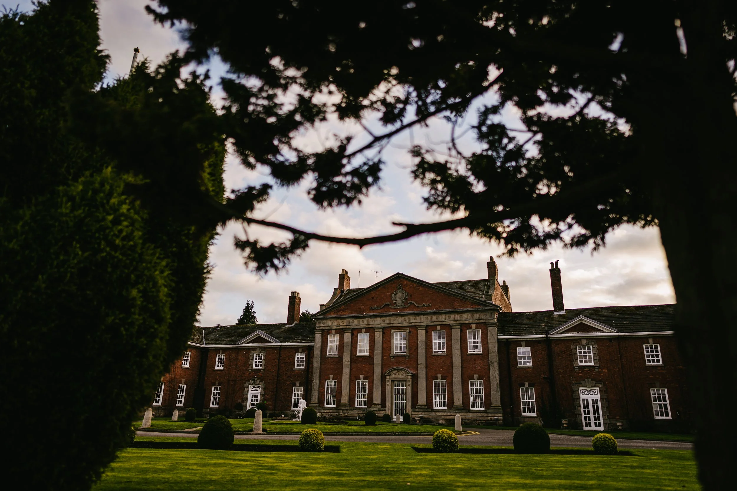A historic brick building with multiple windows and a decorative crest on the roof, surrounded by a well-maintained garden with trimmed bushes and a lawn, viewed through a foreground of trees.