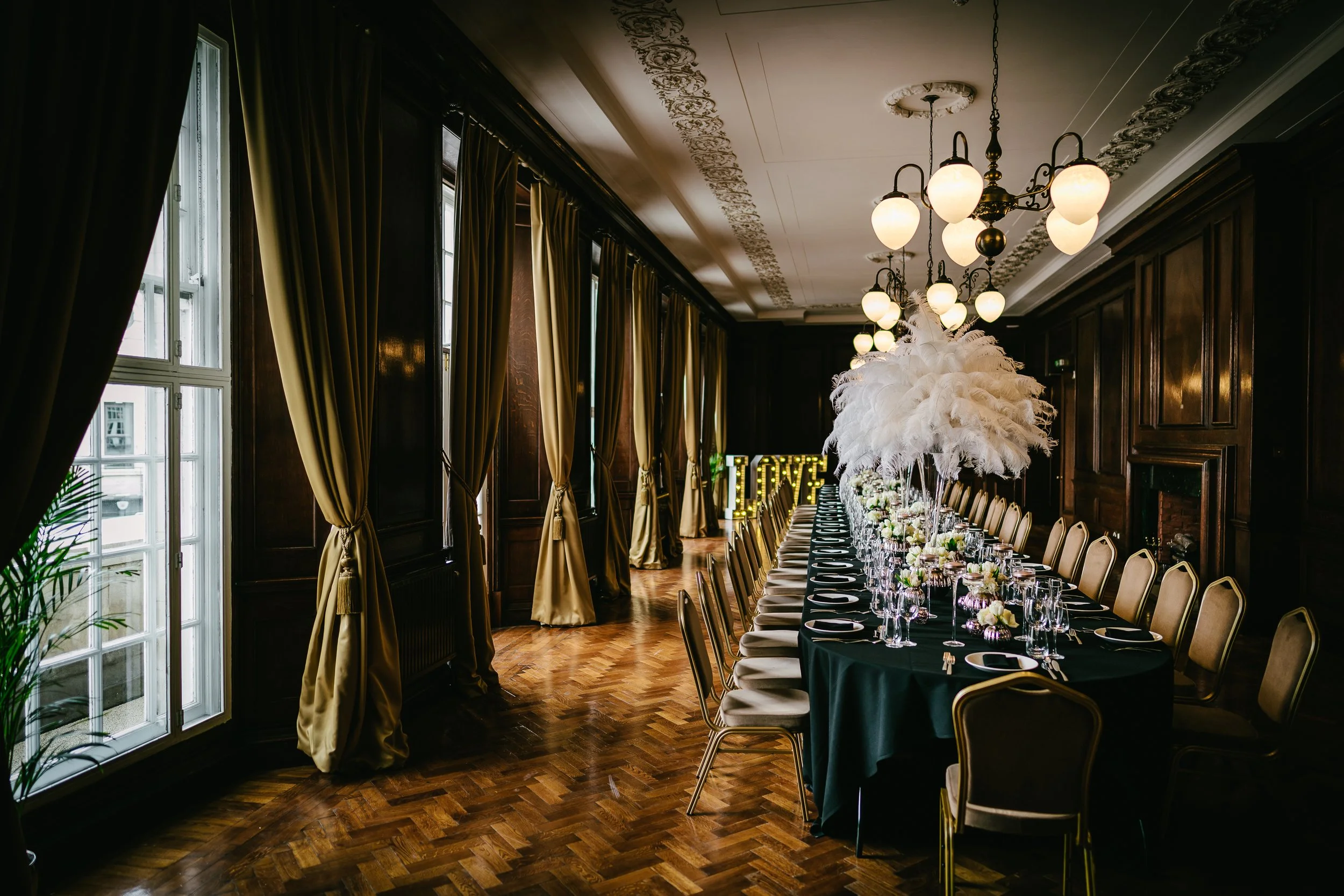 Elegant banquet hall with a long, decorated table set with glasses, plates, cutlery, and floral centerpieces. The room has dark wood paneling, tall windows with gold curtains, and ornate ceiling decorations with warm lighting fixtures. A large illumi