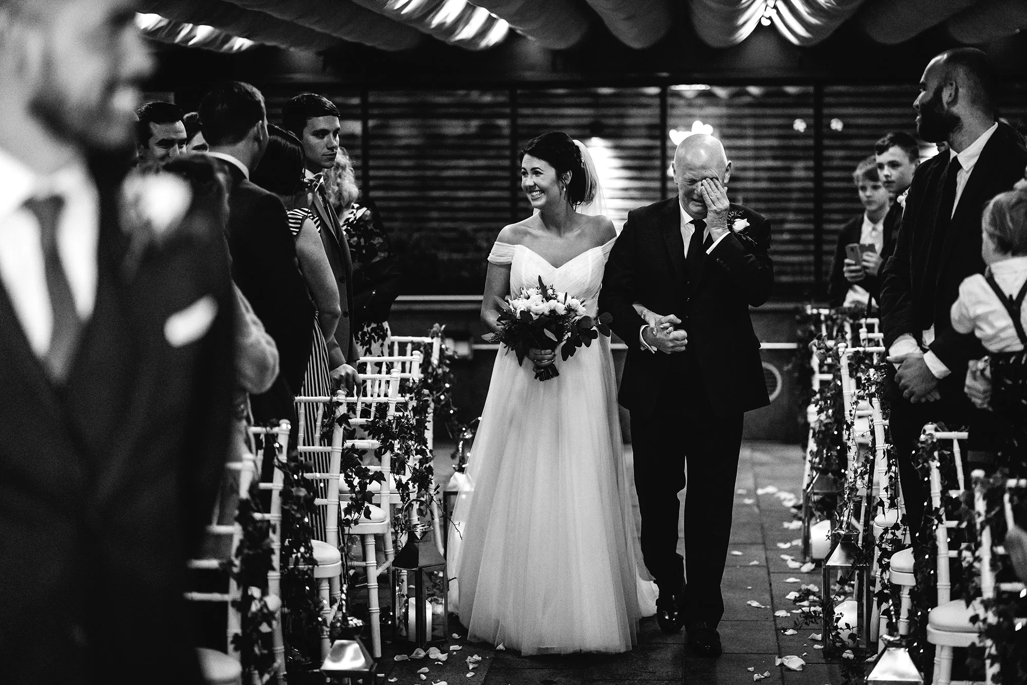 A bride in a wedding dress is walking down the aisle arm-in-arm with an older man, who is wiping a tear from his eye, at a wedding ceremony. Guests are standing on either side of the aisle watching and smiling. The scene is indoors with decorative li
