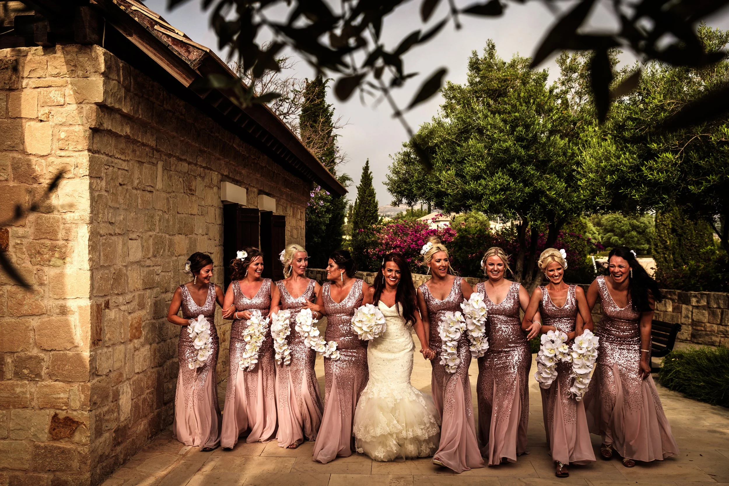 A bride and nine bridesmaids holding hands outdoors against a stone wall, with trees and pink flowering bushes in the background, during a wedding celebration.