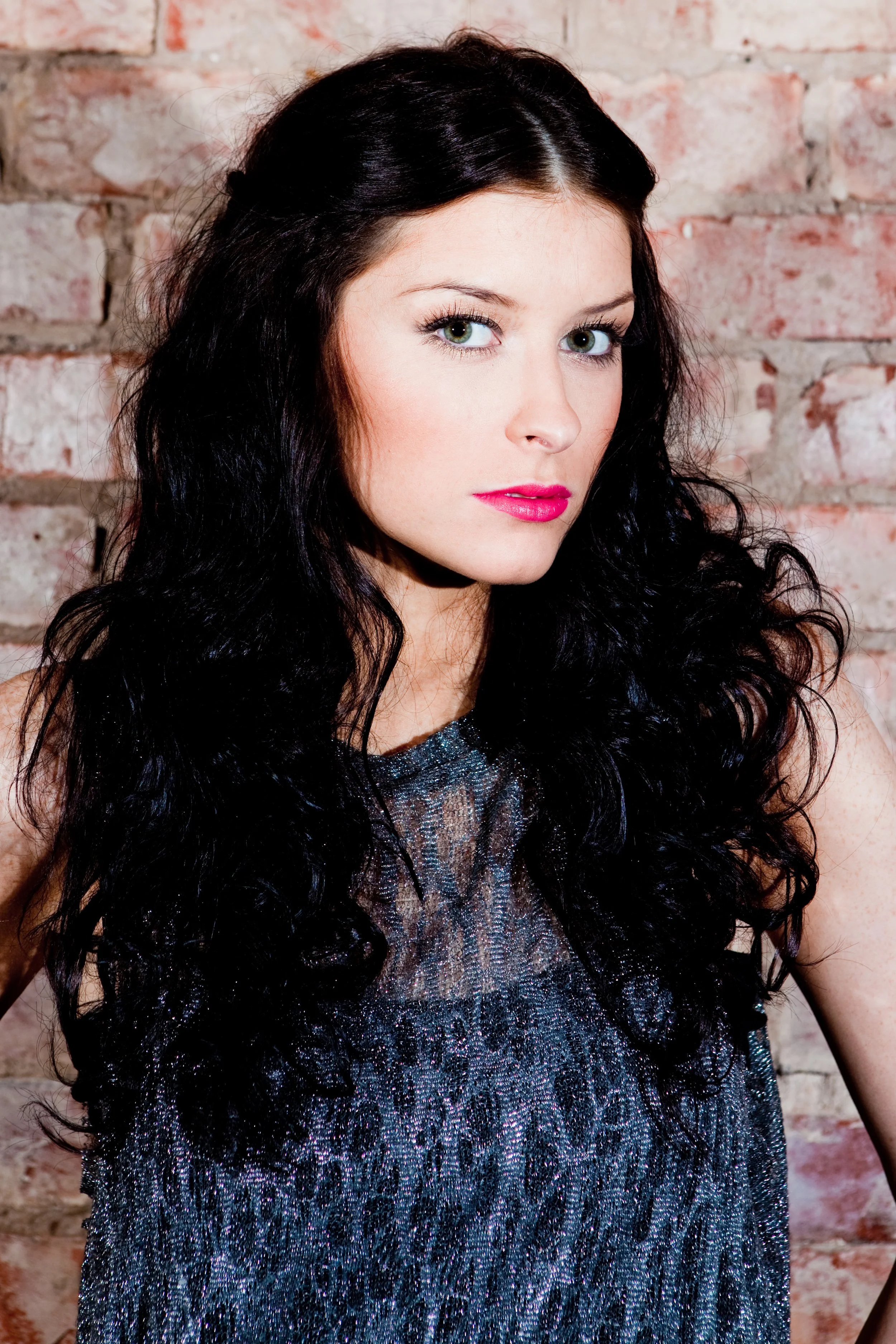 Close-up of a woman with black curly hair, wearing a sparkly dress, against a brick wall background.