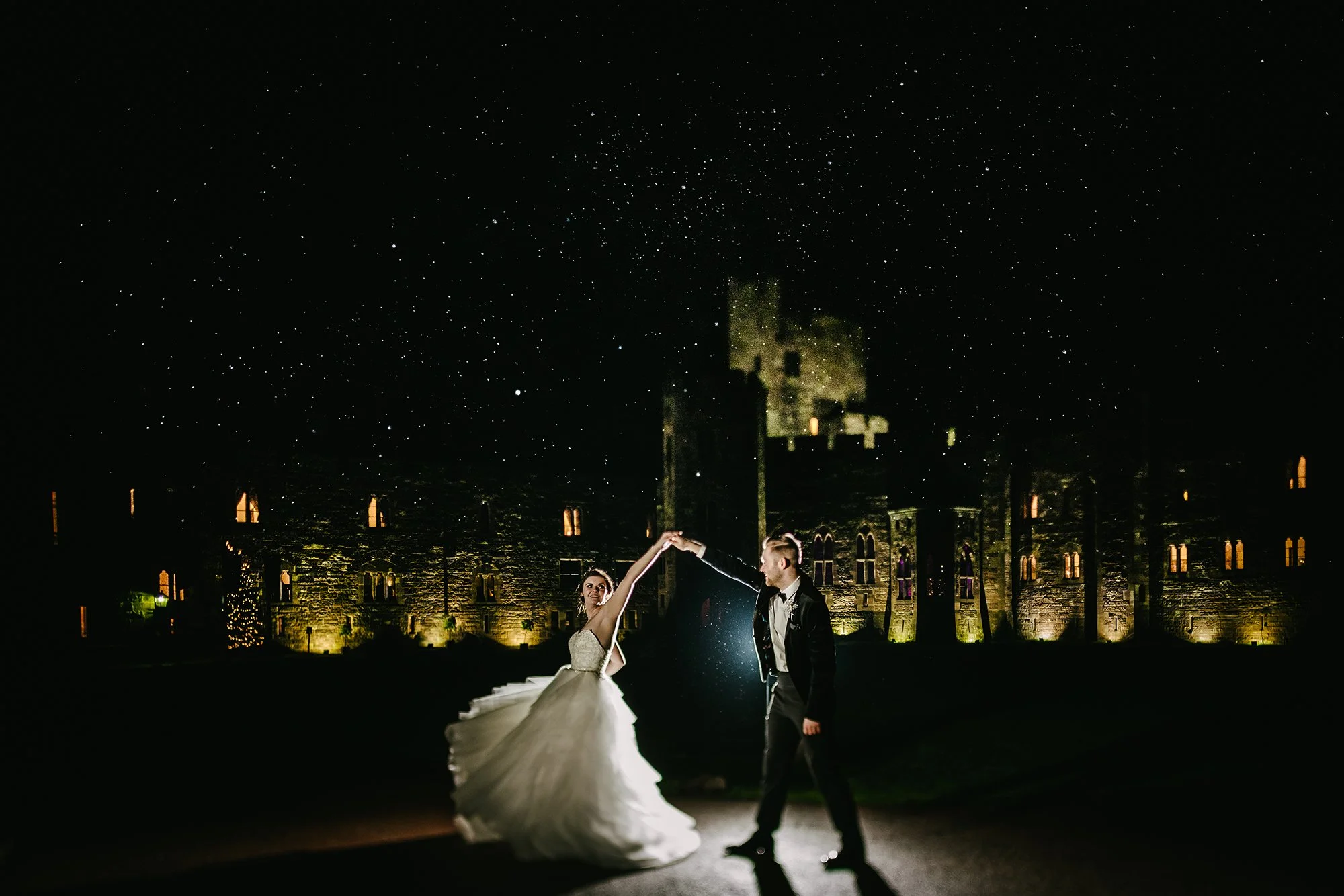 A bride and groom dancing outdoors at night, with a starry sky and historic castle in the background, illuminated by warm lights.