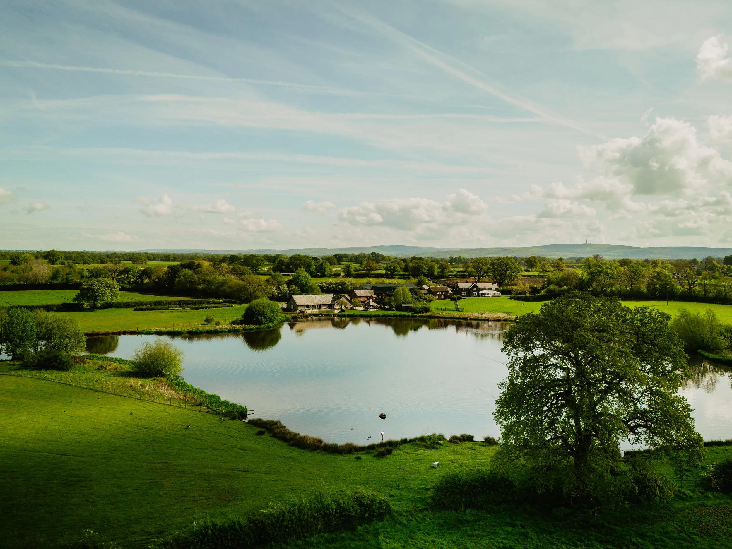 A peaceful countryside landscape with a river, green fields, trees, and houses under a partly cloudy sky.