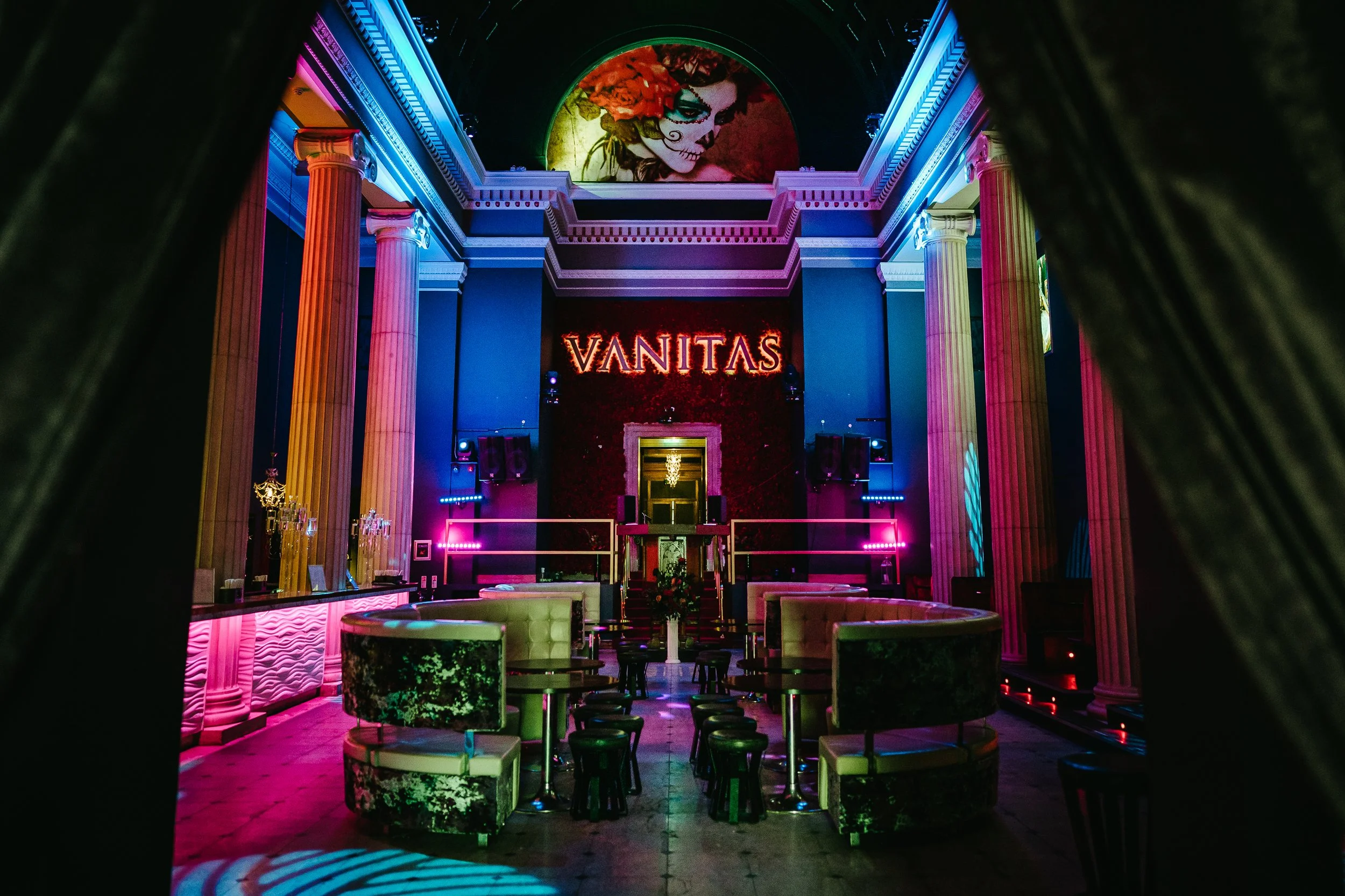 Interior of a nightclub or bar with colorful lighting, tall columns, a dance floor, and a large projection of a woman with painted face and floral headpiece, along with the word 'VANITAS' illuminated on the wall.