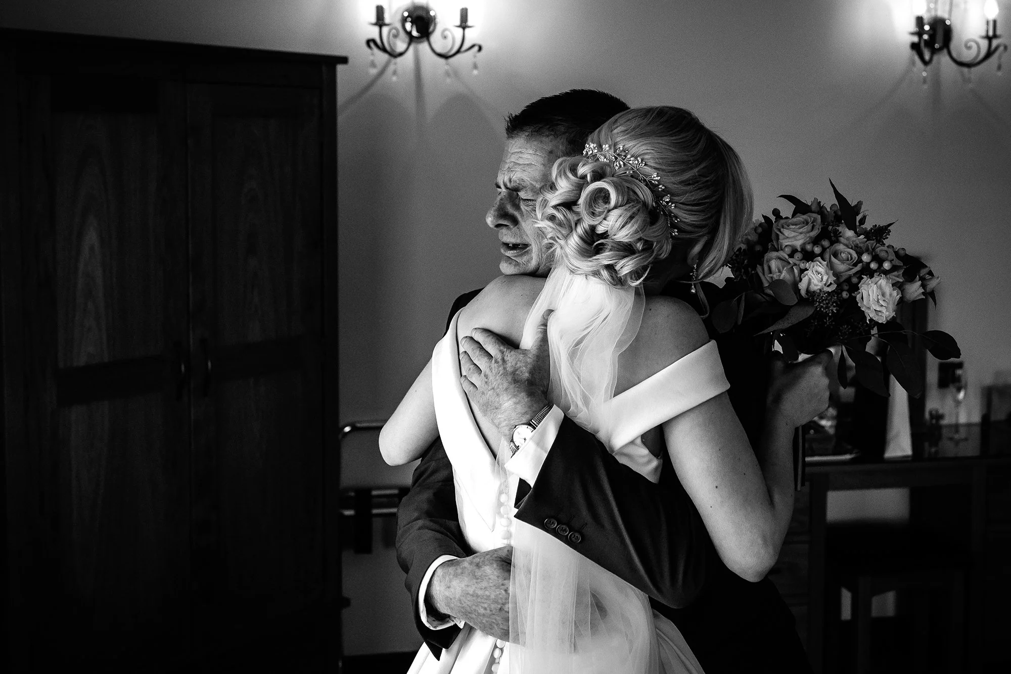 A bride hugging an older man, likely her father, during a wedding in an indoor setting. The bride is holding a bouquet and has styled blonde hair with a headpiece. The scene is in black and white.