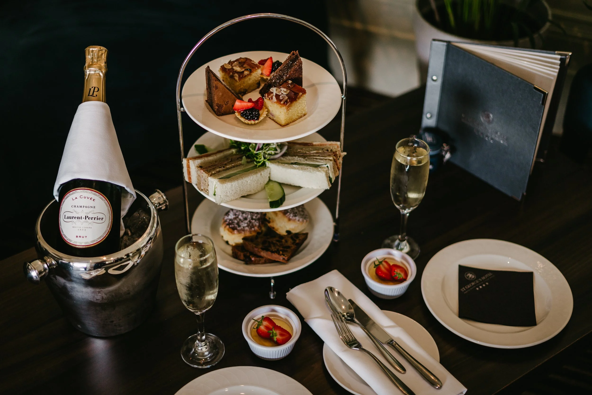 A multi-tiered serving tray with various cakes and desserts, a bottle of Laurent-Perrier champagne in an ice bucket, two glasses of champagne, and small bowls of strawberries on a dark table in a fine dining setting.