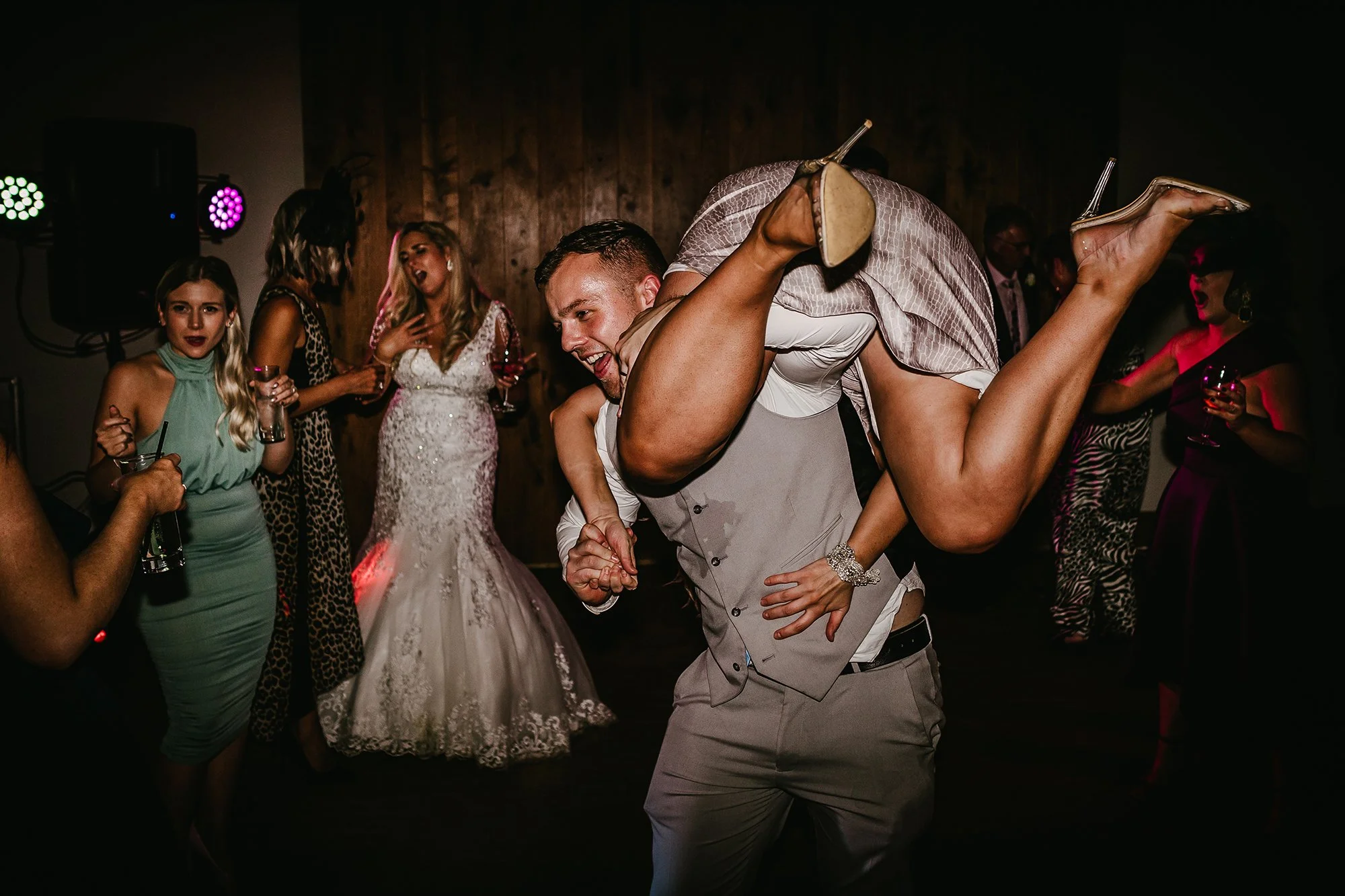 A groom dances with a woman on a wedding reception dance floor, surrounded by guests, some holding drinks, with a wooden wall in the background and colorful party lighting.