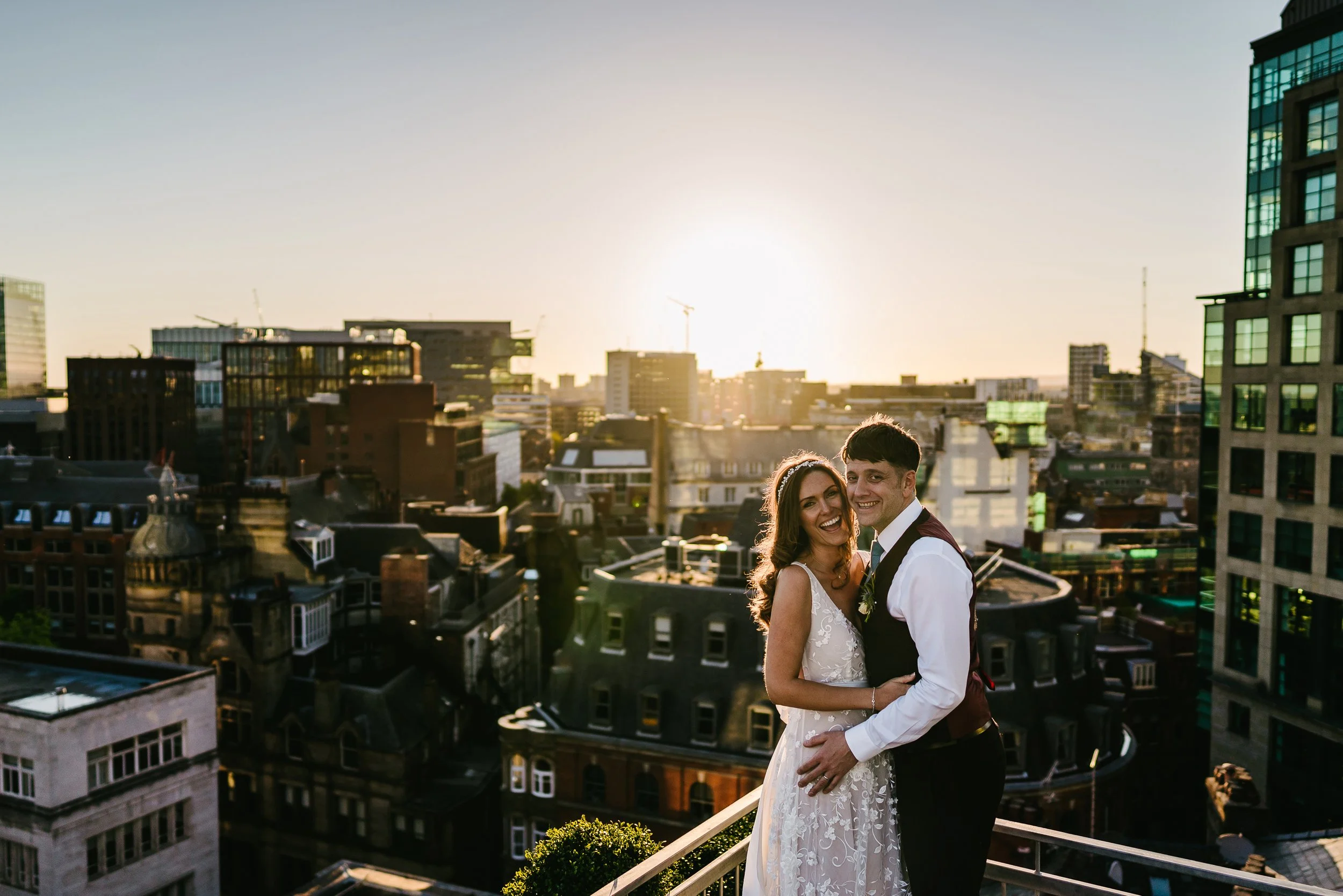 A newly married couple standing on a balcony with a city skyline during sunset, smiling and embracing.