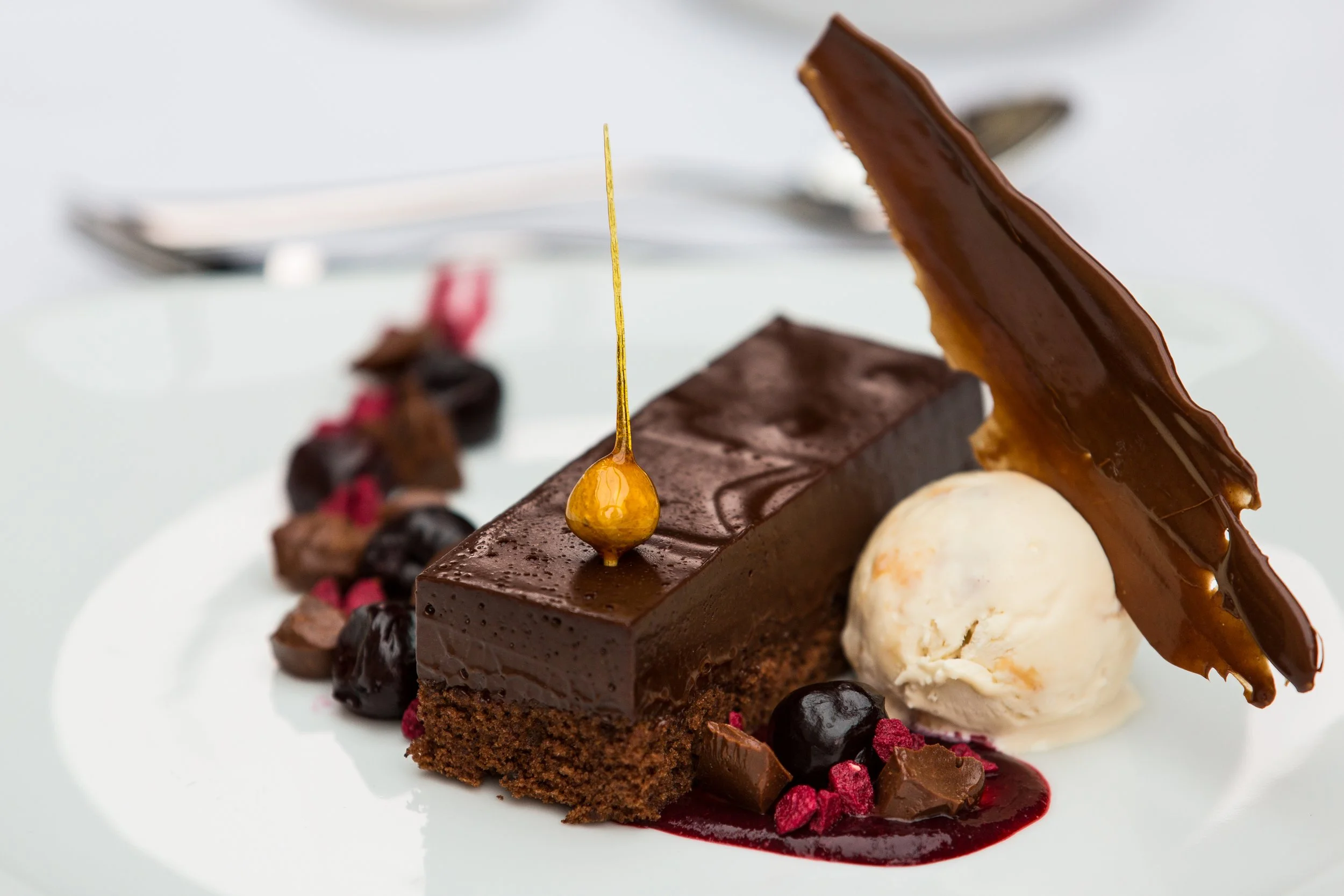 Close-up of a chocolate dessert on a white plate, with a square piece of chocolate cake topped with a shiny caramel drizzle, surrounded by berries, sauce, and a dollop of vanilla ice cream.