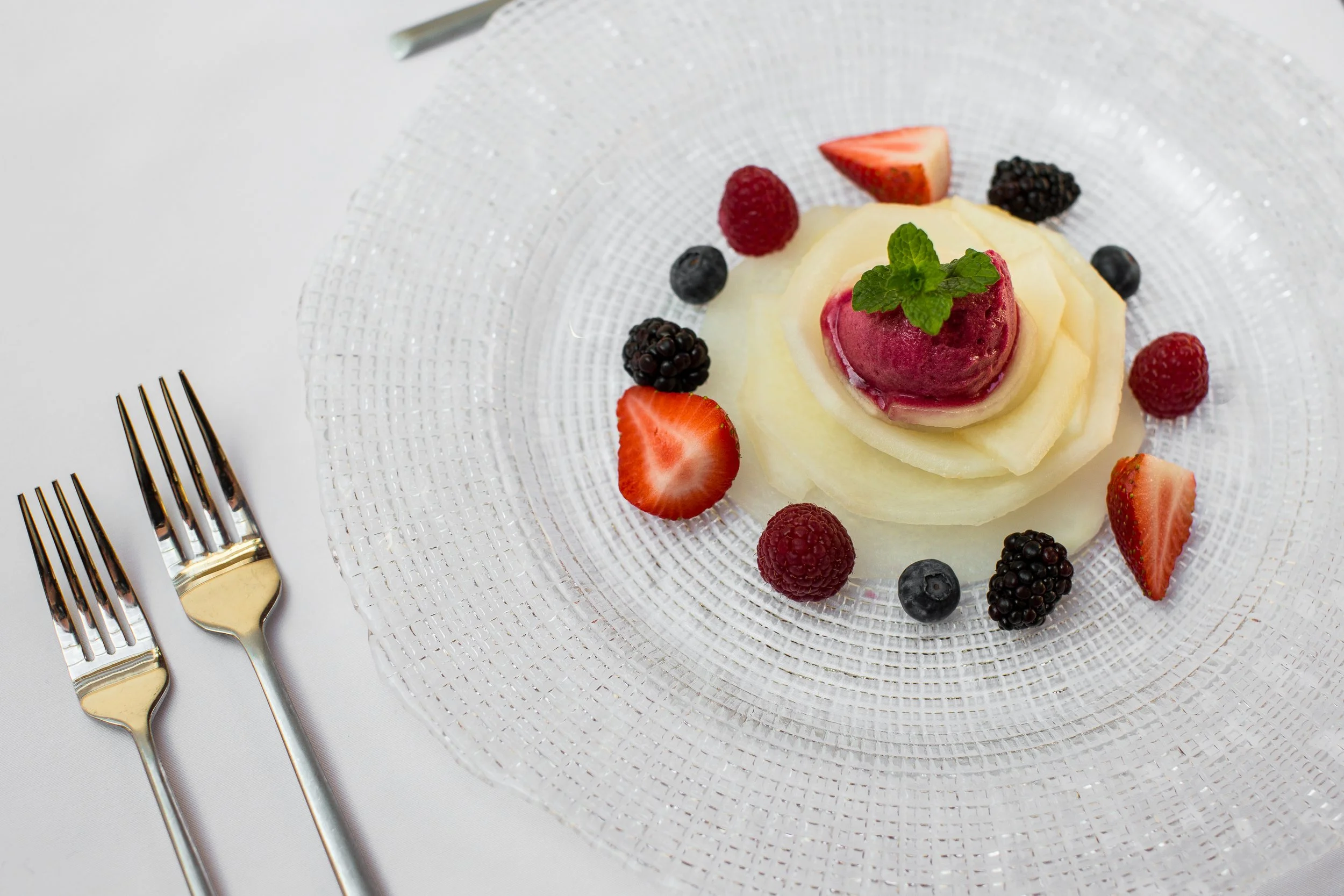 Dessert plate with slices of melon, topped with strawberry sorbet and garnished with a mint sprig, surrounded by fresh berries including strawberries, raspberries, blackberries, and blueberries, on a textured glass plate with gold and silver forks ne