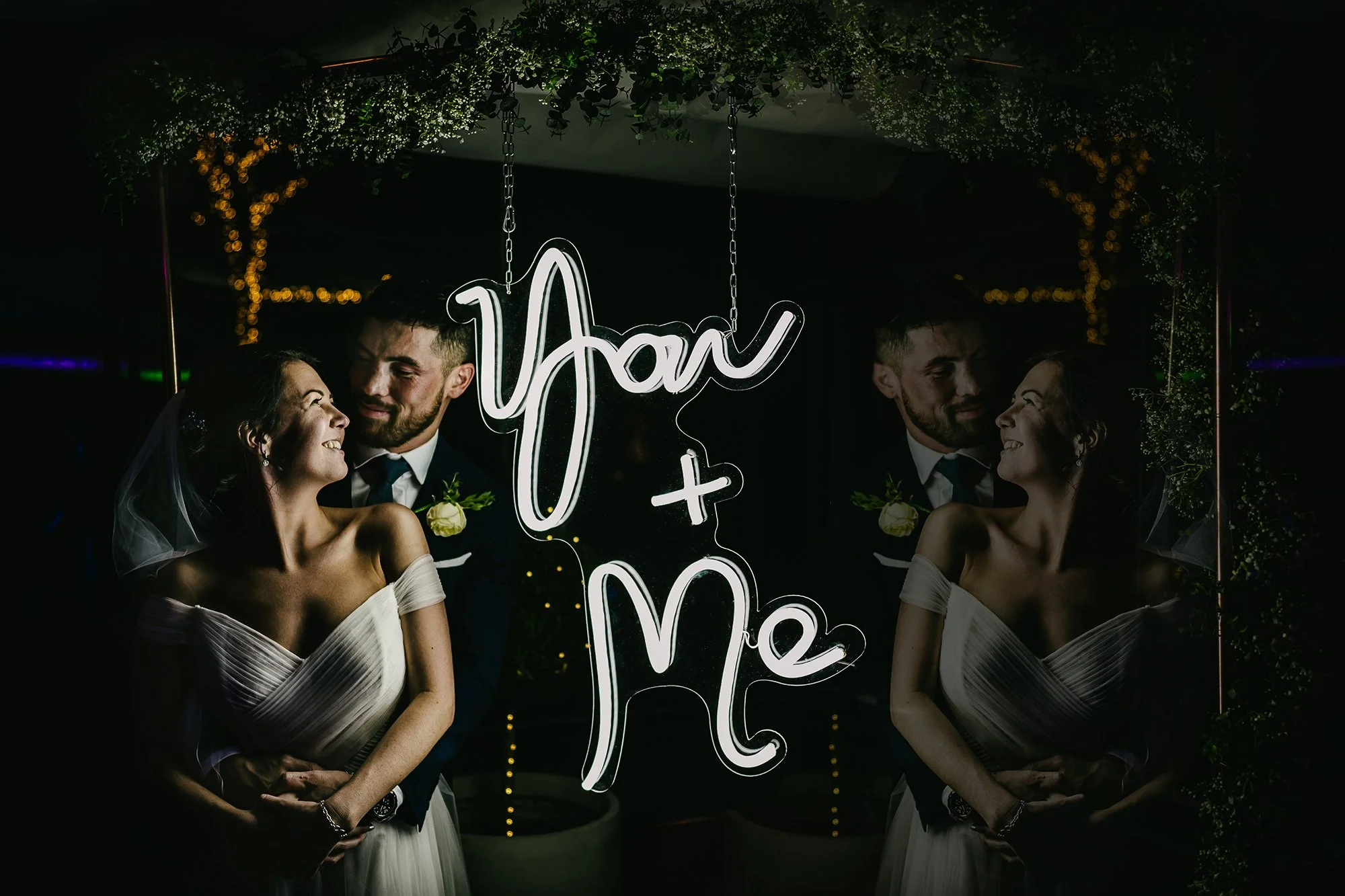 Bride and groom smiling at each other at wedding reception with neon sign reading "You + Me" surrounded by greenery and fairy lights.