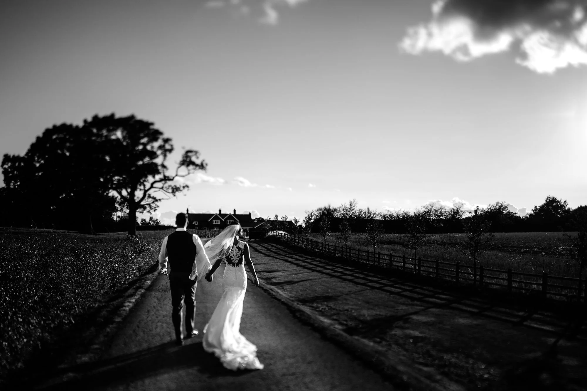 A black and white photograph of a bride and groom walking hand in hand down a rural road, with trees and houses in the background.