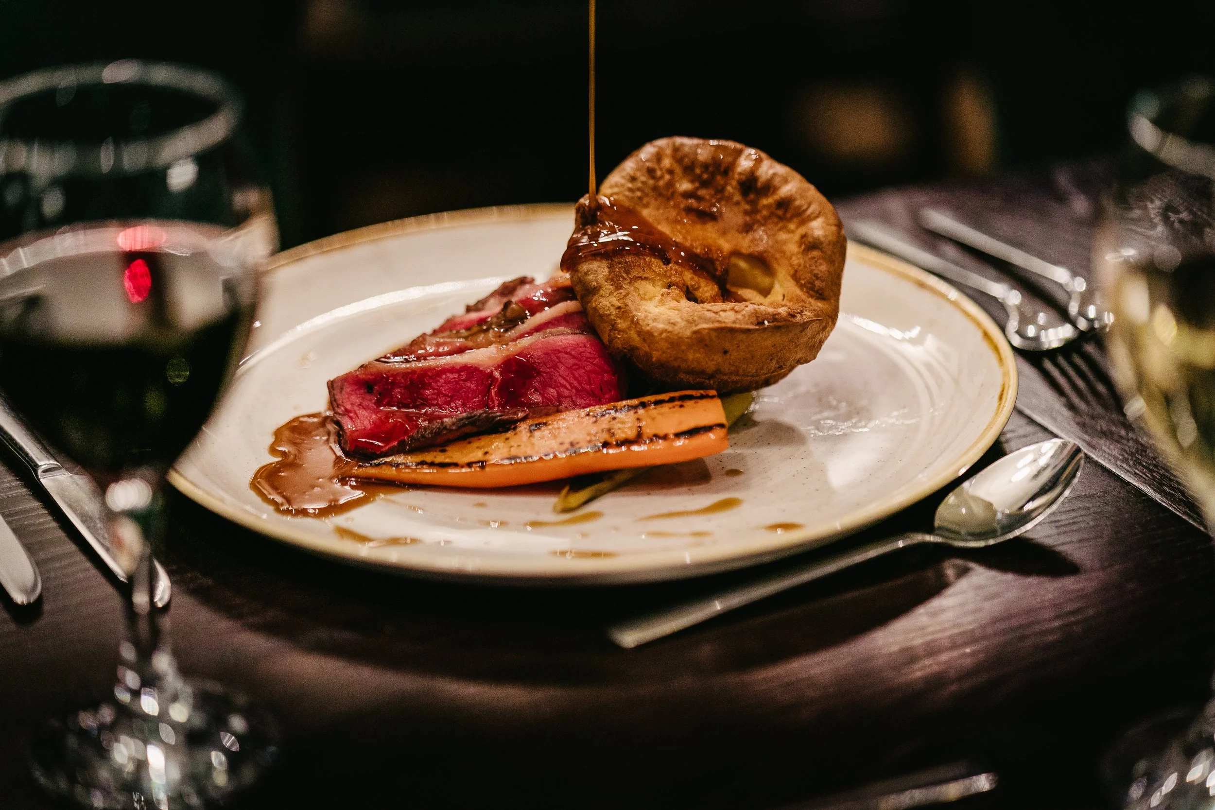 A plated meal with sliced beef, roasted vegetables, a baked potato with butter and gravy, and a drizzle of sauce, on a white plate with a gold rim, set on a dark table.