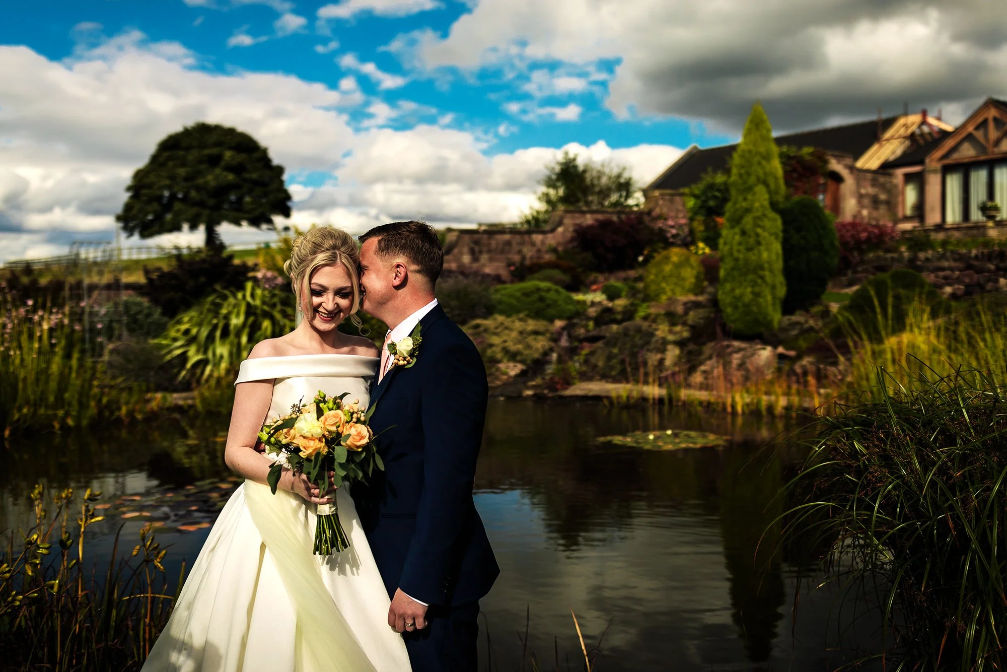 A newlywed couple stands by a pond outdoors, with the bride holding a bouquet of flowers. The groom whispers to the bride's head, both smiling, under a partly cloudy sky.