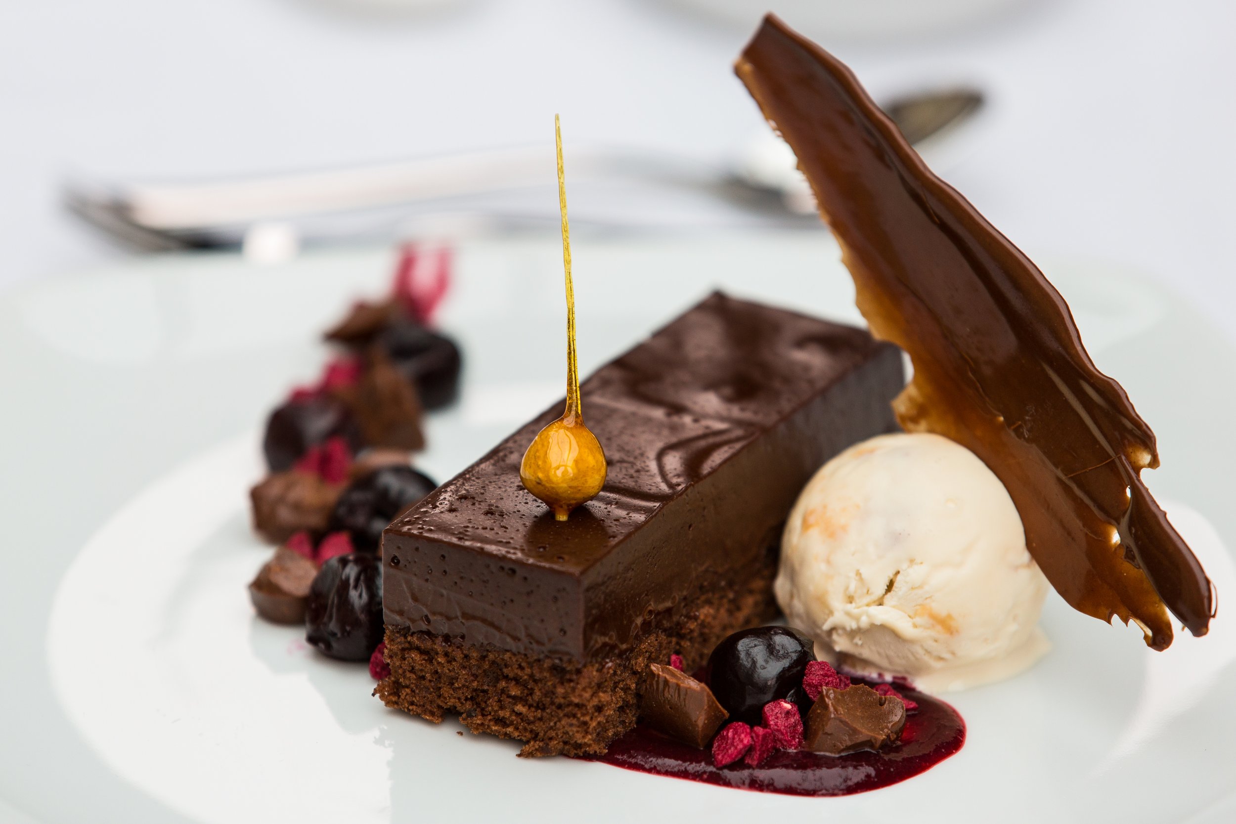 Close-up of a plated chocolate dessert with a rectangular chocolate cake, a slice of caramel-topped chocolate tart, a scoop of vanilla ice cream, and fruit garnishes on a white plate.