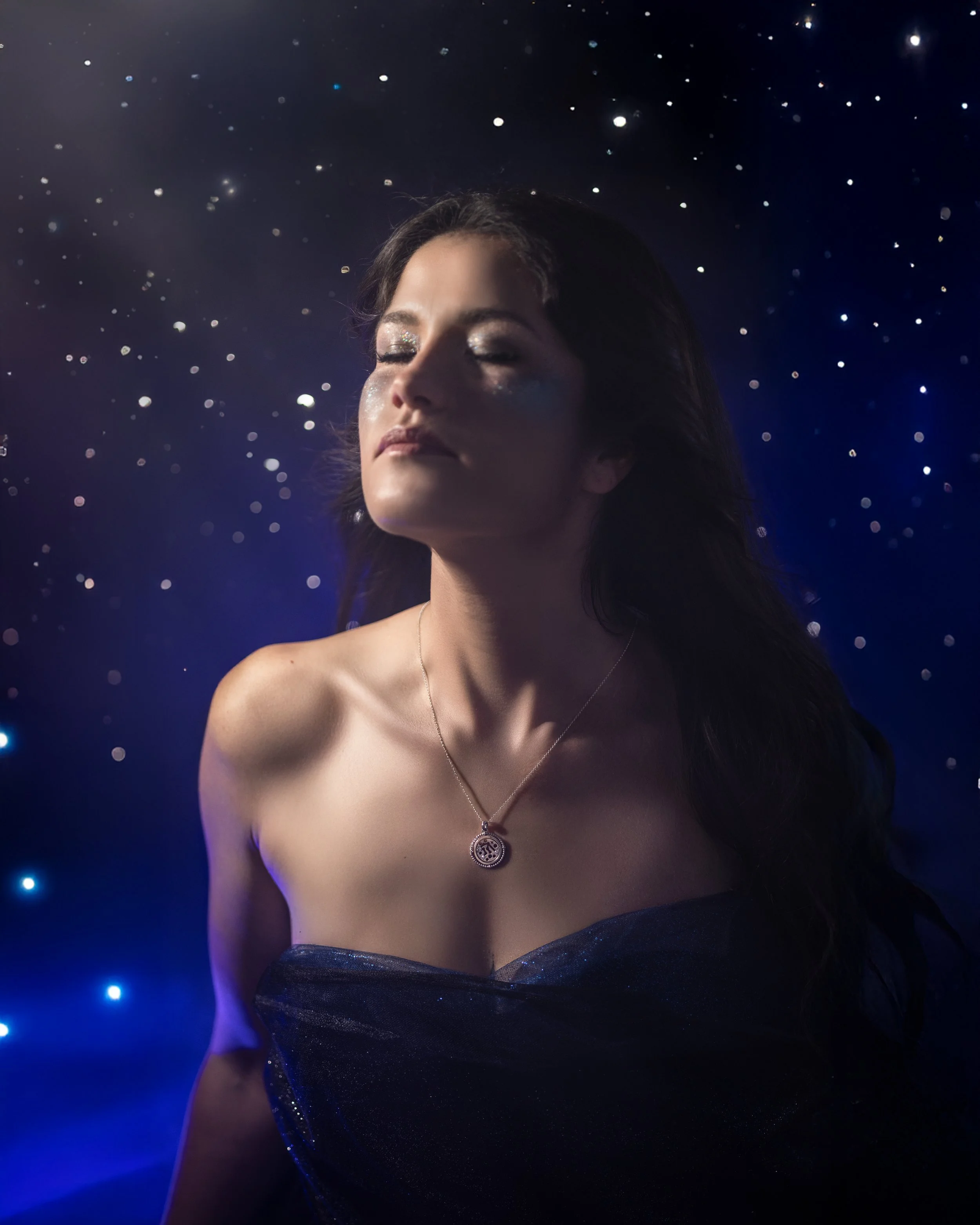 Femme avec les yeux fermés, portant un collier avec un pendentif, vêtue d'une robe noire scintillante, devant un fond sombre avec des lumières bleues et blanches.