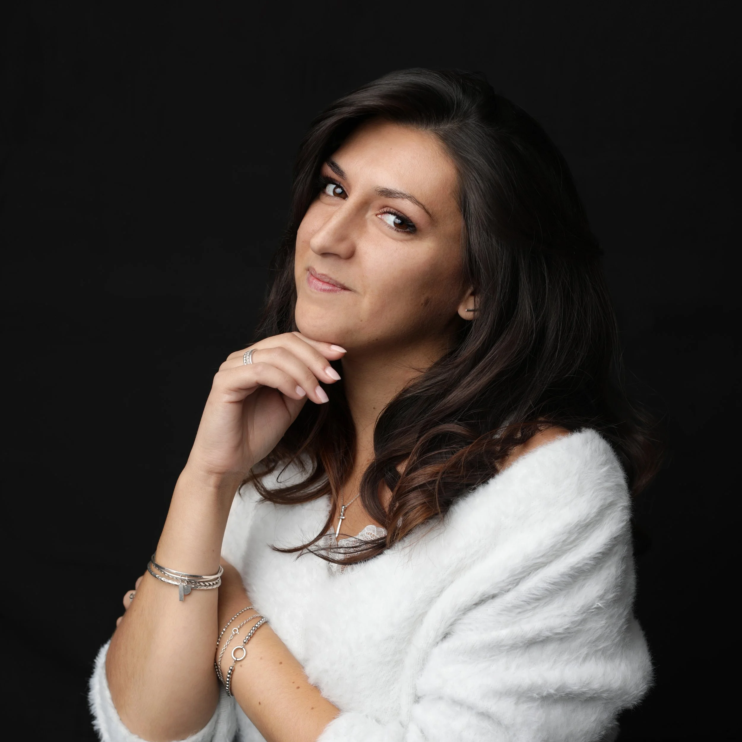 Portrait de la créatrice de la marque ael bijoux avec des cheveux bruns, portant un pull blanc, des bijoux en argent, posant avec la main sous le menton contre un fond noir.