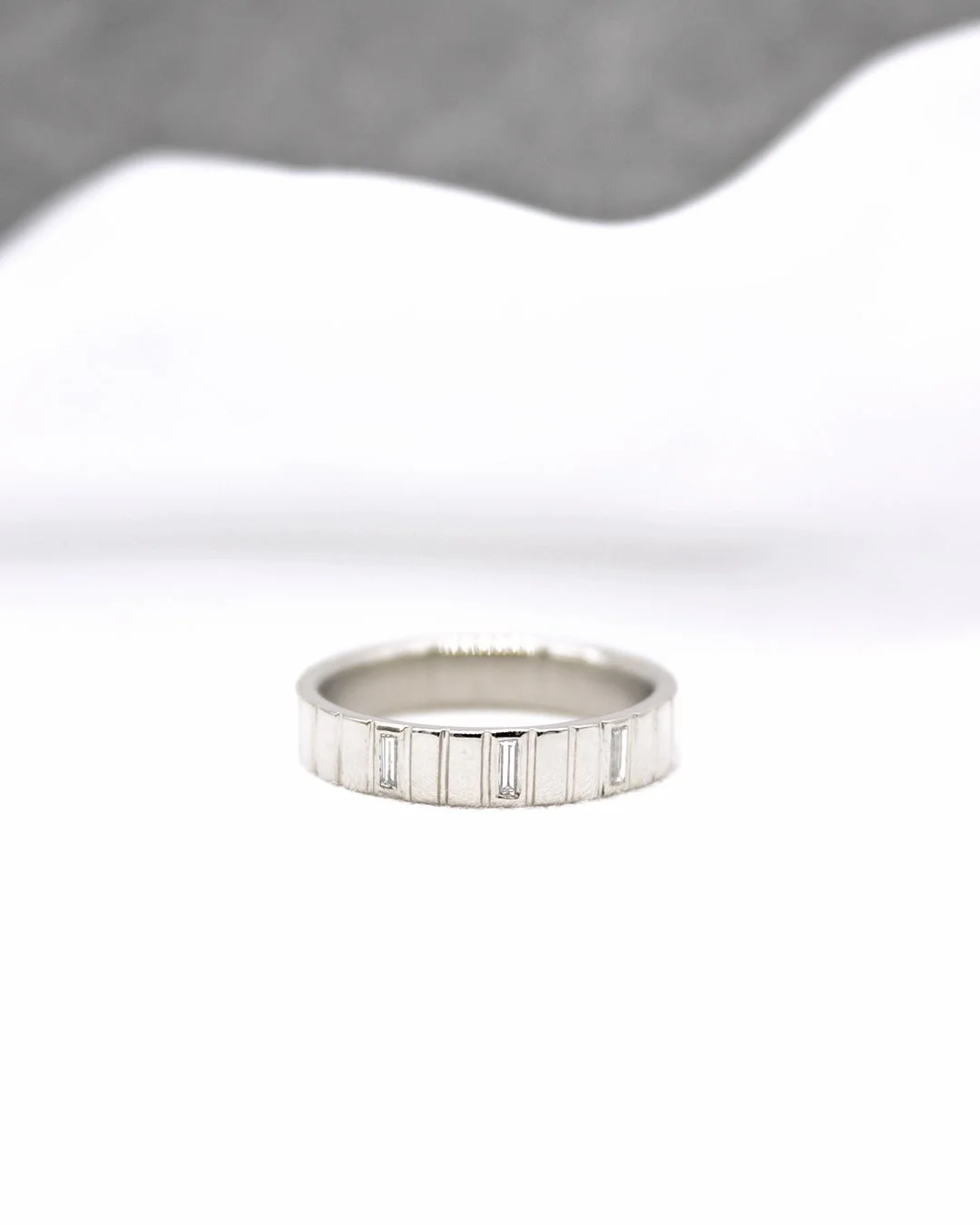 Bague en argent avec des diamants rectangulaires, posée sur une surface blanche, avec des tissus gris et blanc en arrière-plan.
