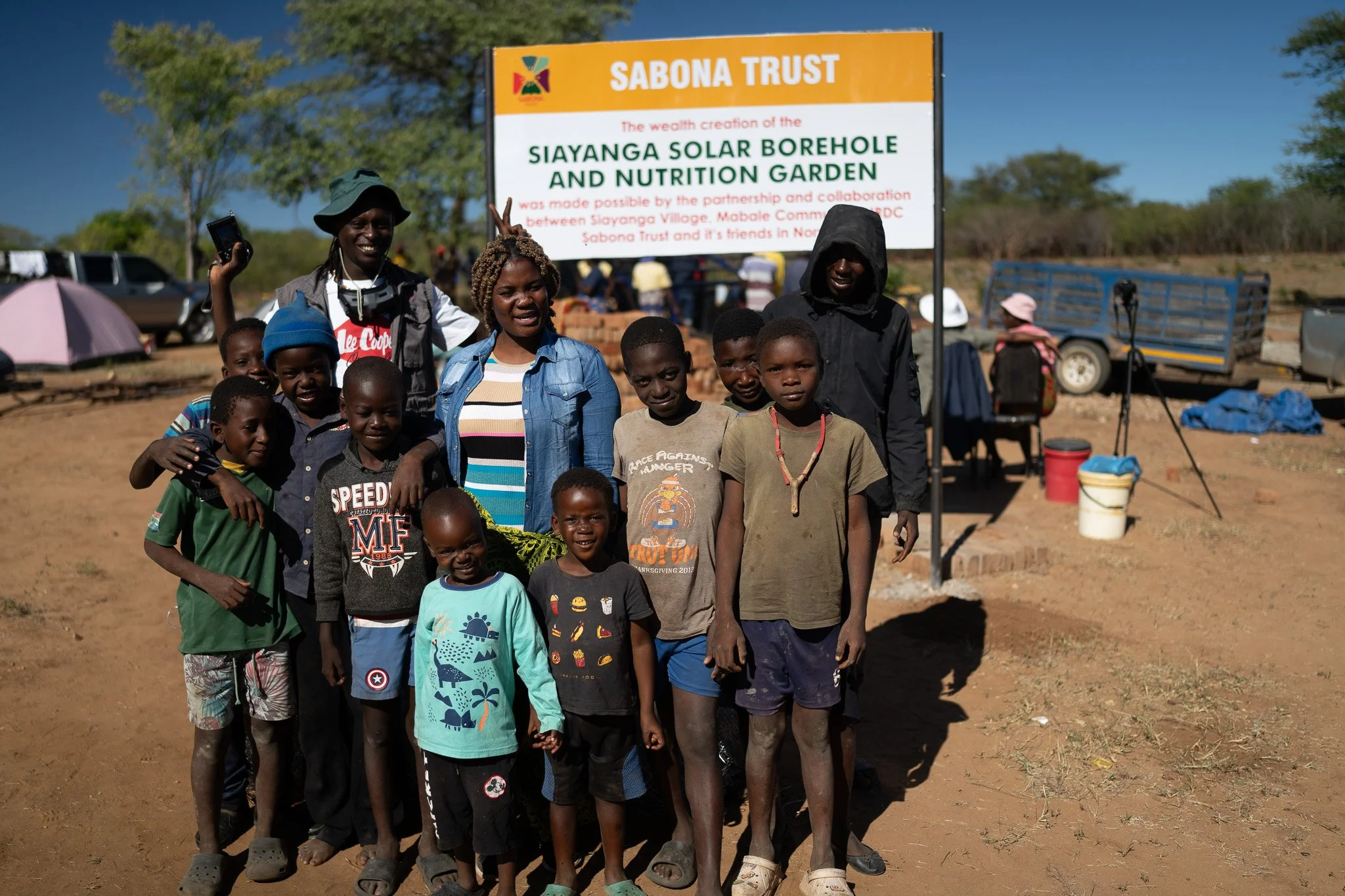 En gruppe mennesker står foran en stor skilting som sier 'SIAYANGA SOLAR BOREHOLE AND NUTRITION GARDEN', med flere barn og voksne smilende, utendørs i et tørt landskap med trær i bakgrunnen.