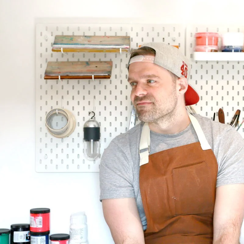 Man with a beard wearing a gray t-shirt, a backwards cap, and an apron in a workshop or craft room.