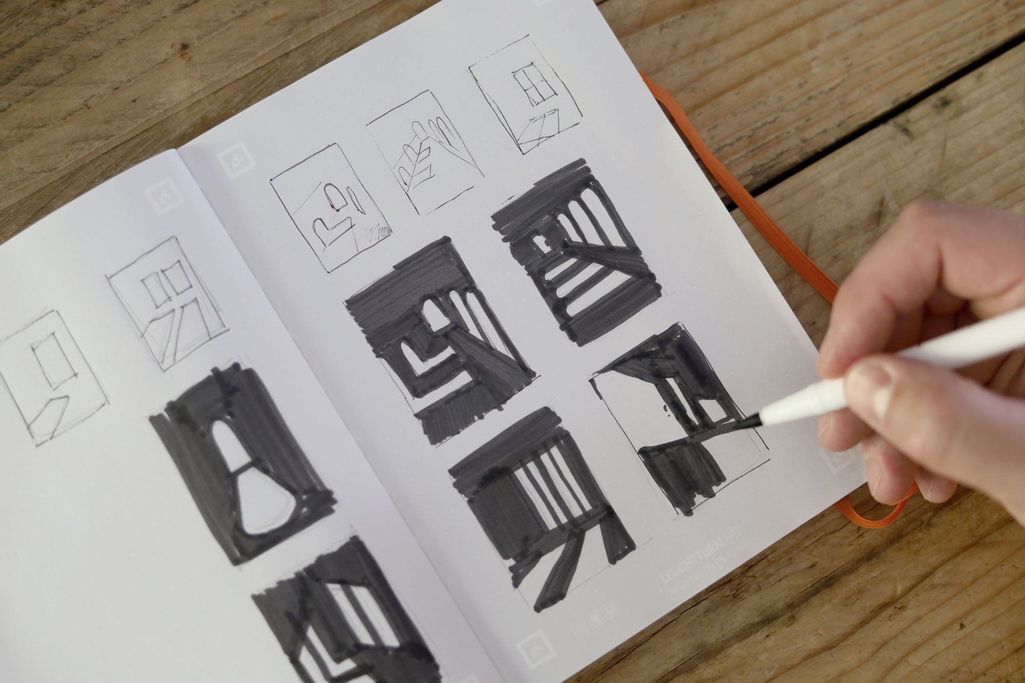 Sketches of window designs and black paint or ink blocks on a sheet of paper, being held and drawn by a person's hand with a white pen, placed on a wooden surface.
