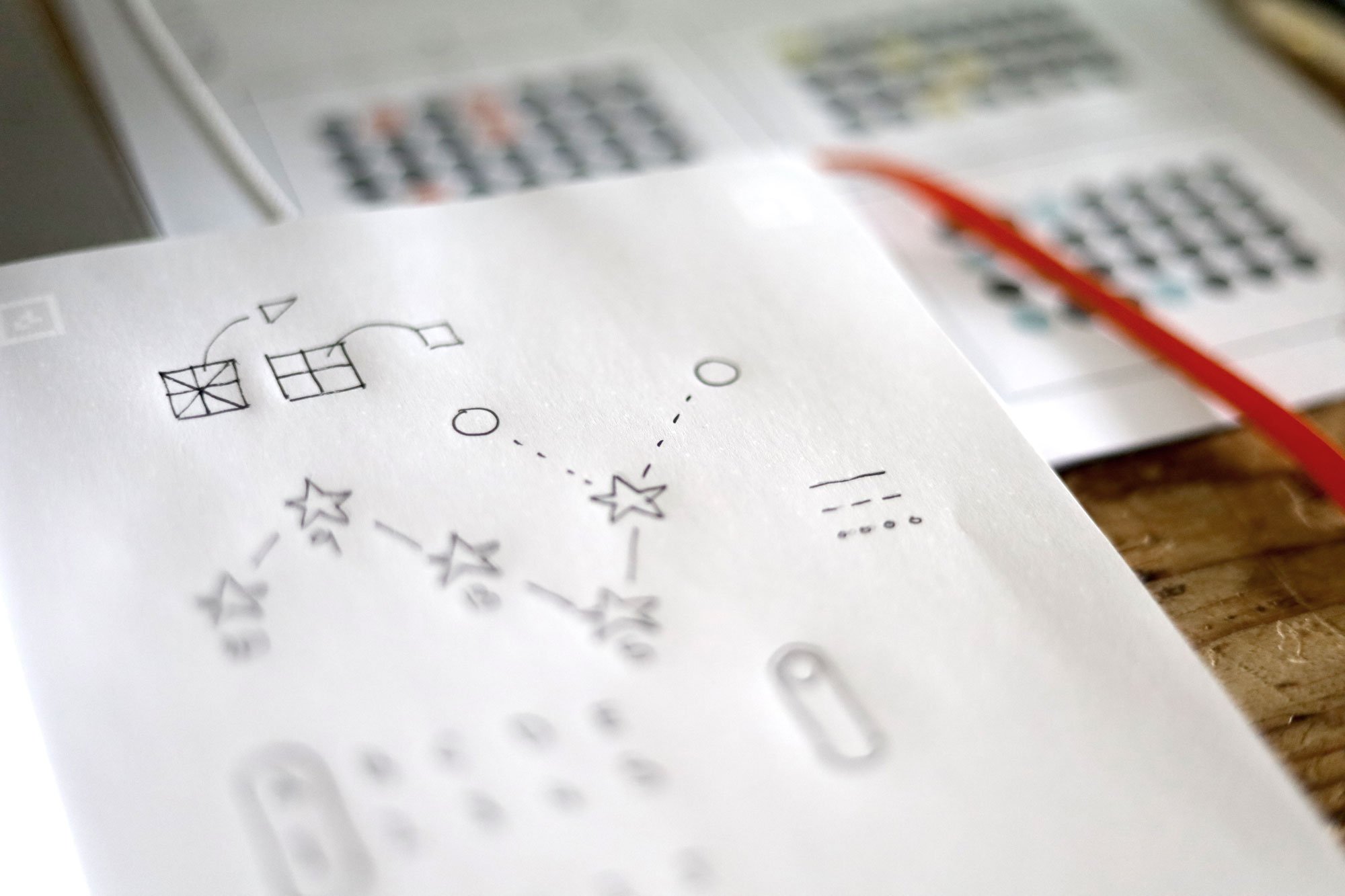 Close-up of a piece of paper with hand-drawn shapes and dotted lines, on a wooden surface. Blurred background with patterned objects.
