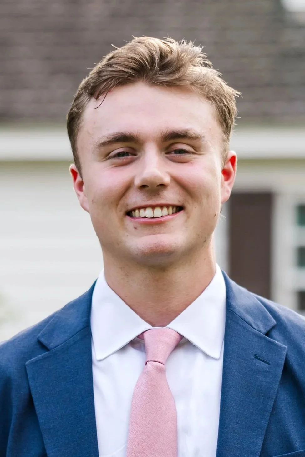 Portrait of a young man in a blue suit and pink tie, smiling outdoors with a house in the background.