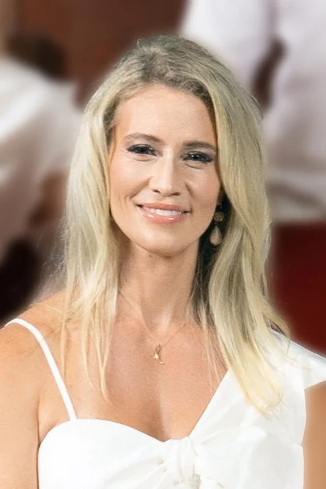 A woman with long blonde hair, smiling, wearing a white dress with spaghetti straps and a small necklace, standing indoors with blurred background.