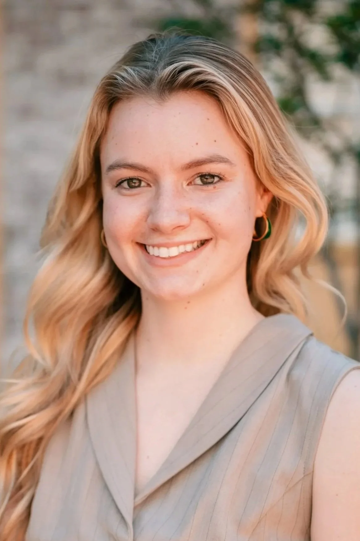 A young woman smiling outdoors with long, wavy blonde hair, wearing a sleeveless beige top and gold hoop earrings.