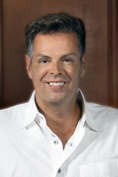 Portrait of a man with dark hair smiling, wearing a white shirt, with a wooden background.