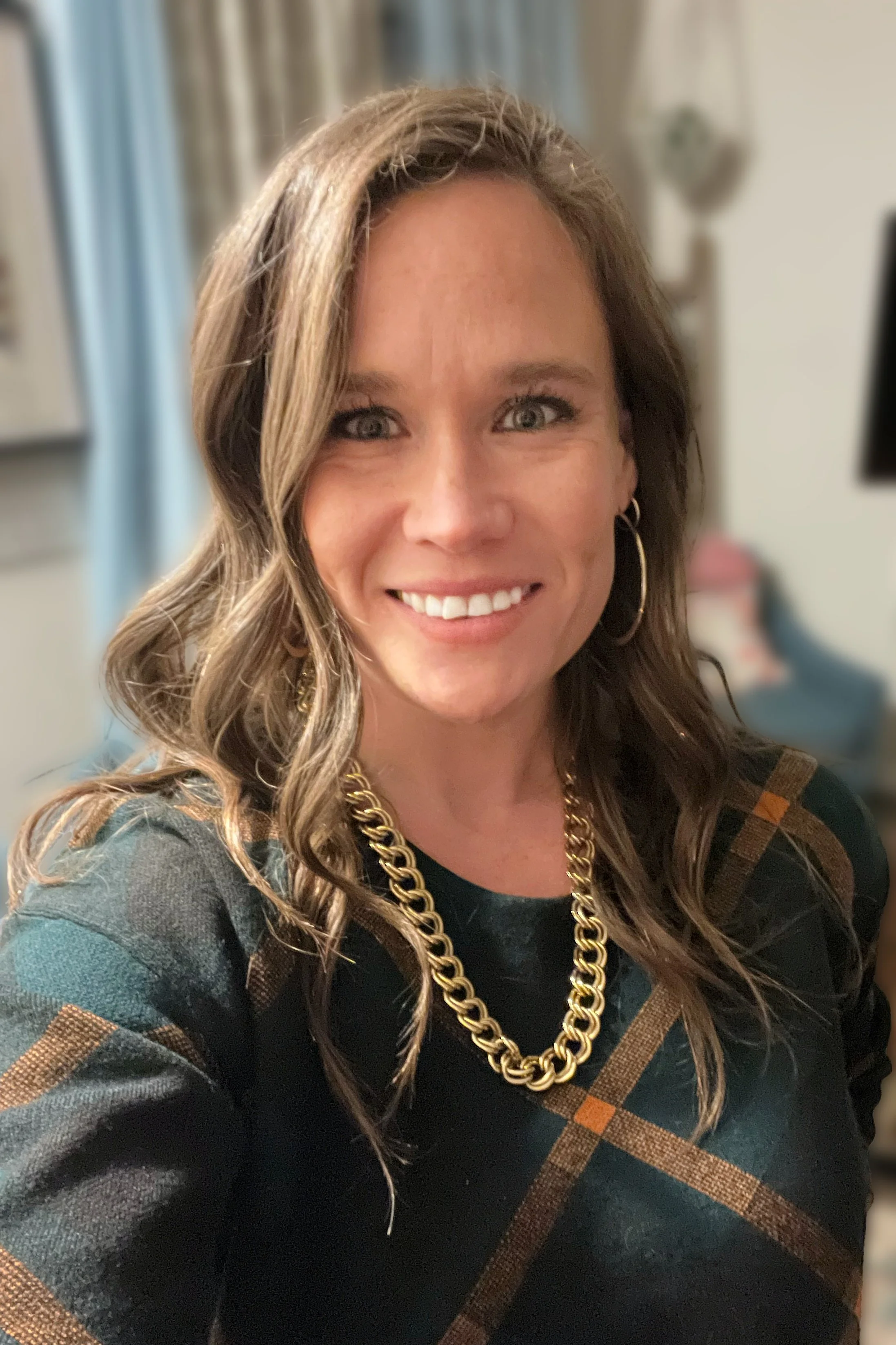 A woman smiling, wearing a dark plaid shirt with gold jewelry, including hoop earrings and a chain necklace, in a cozy room.