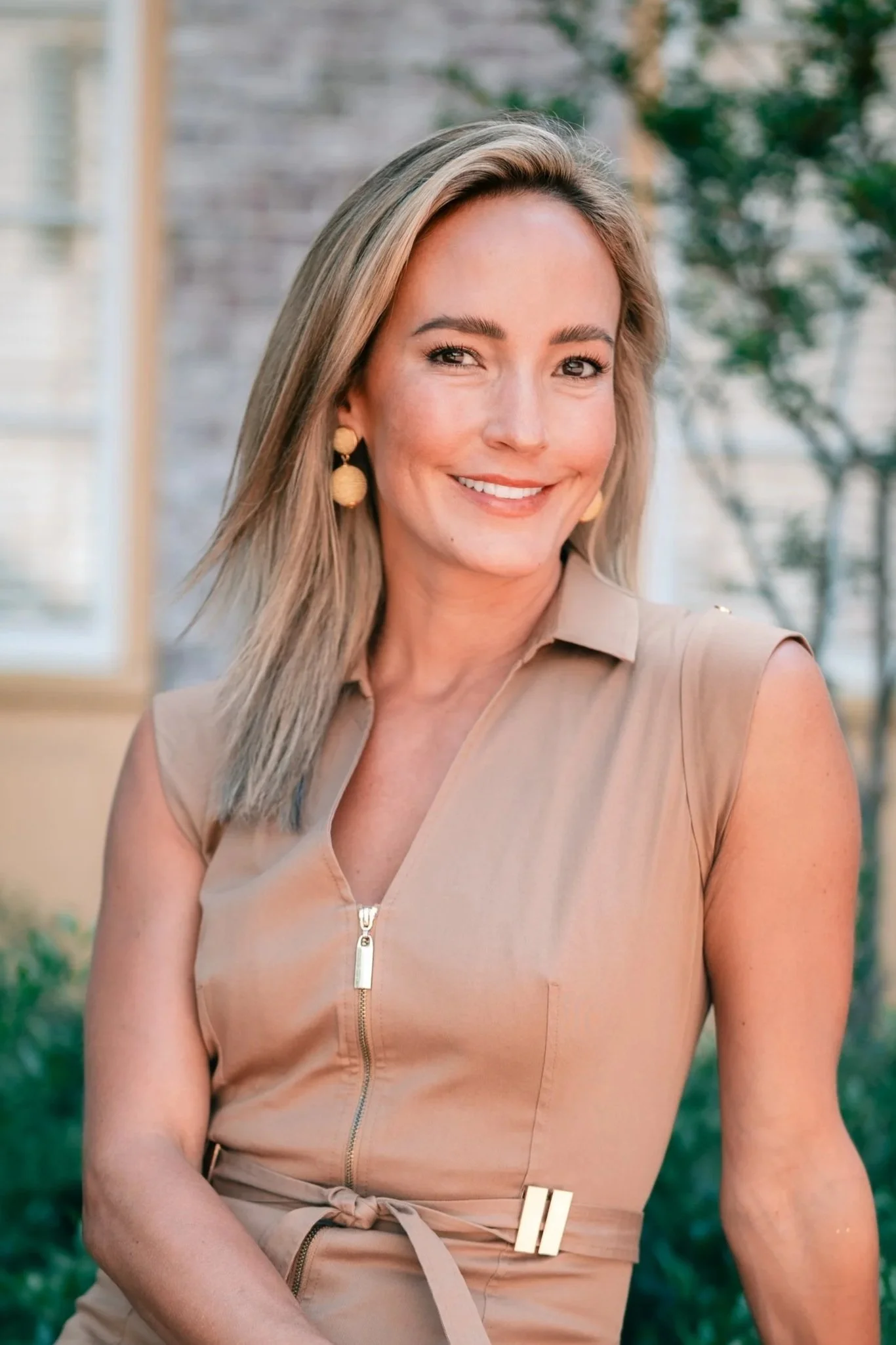 A woman smiling outdoors, wearing a sleeveless beige dress with a zipper and belt, with trees and a house in the background.