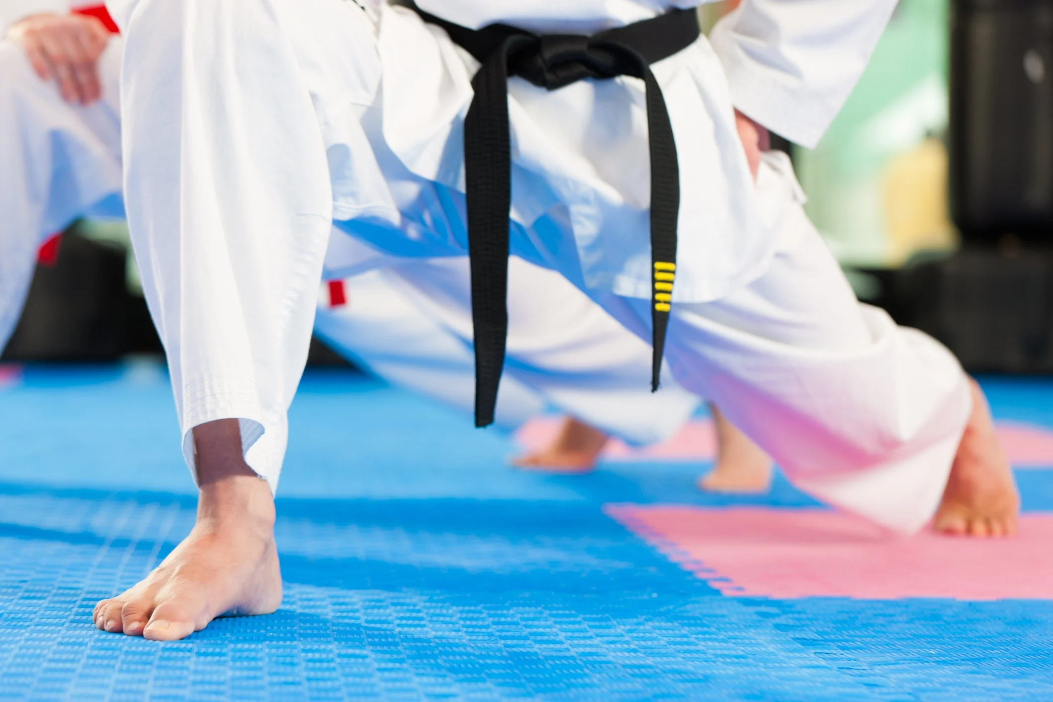 Close-up of martial artist's legs in a lunge stance wearing a white gi and black belt on a blue and pink mat.
