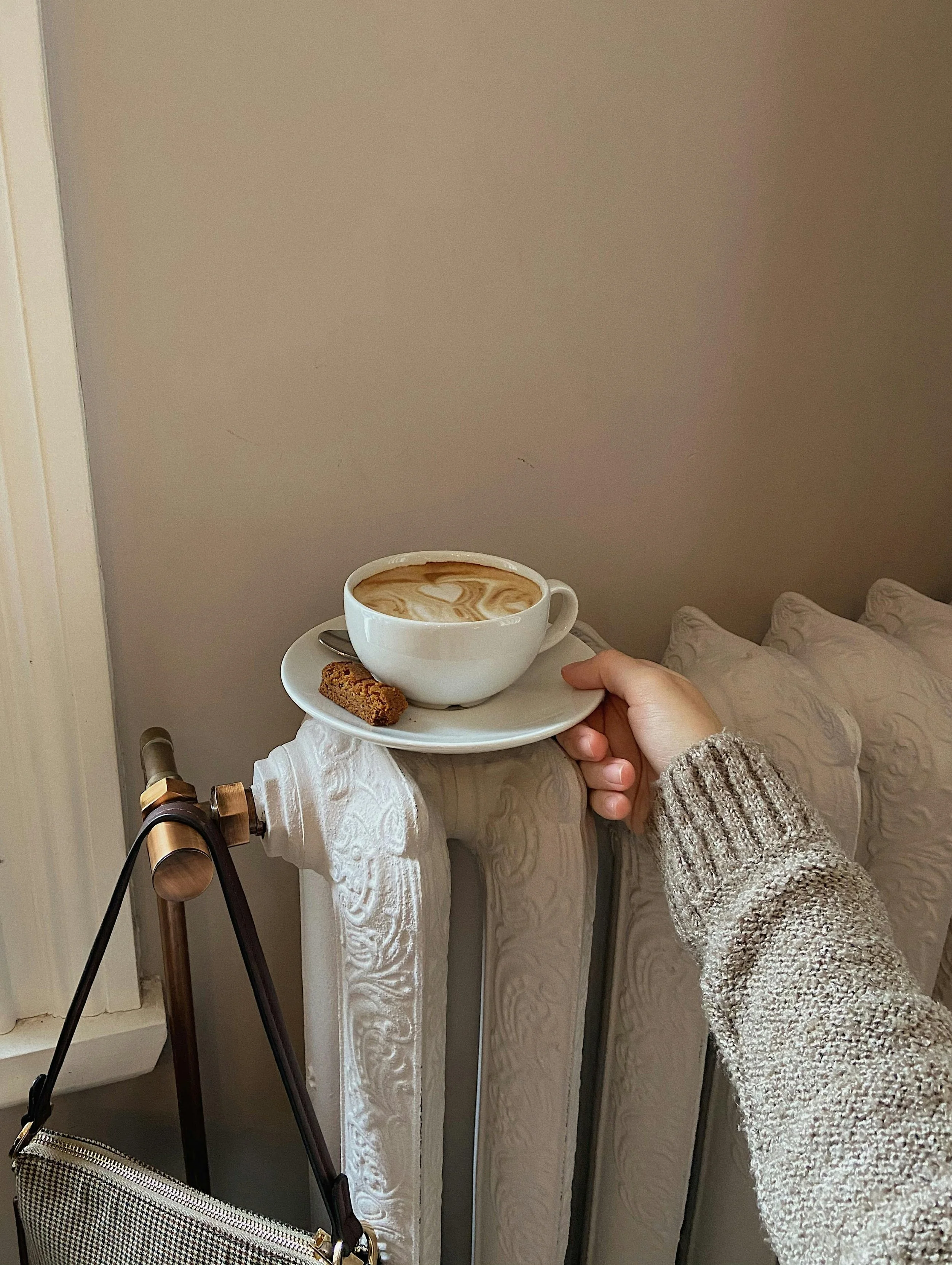 A person in a gray, textured sweater holding a white tray with a cup of coffee with latte art and two cookies, in a room with a gray wall and a bed with cream-colored bedding.