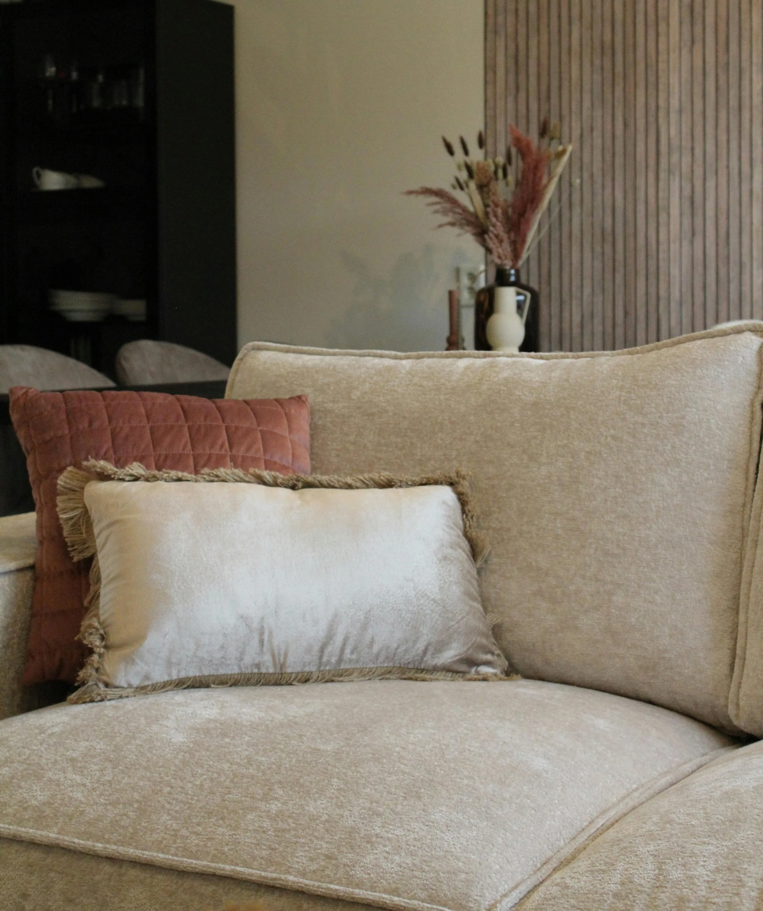 Living room with beige sofas, decorative pillows, and a vase with dried flowers on a table against a wall with wood paneling.