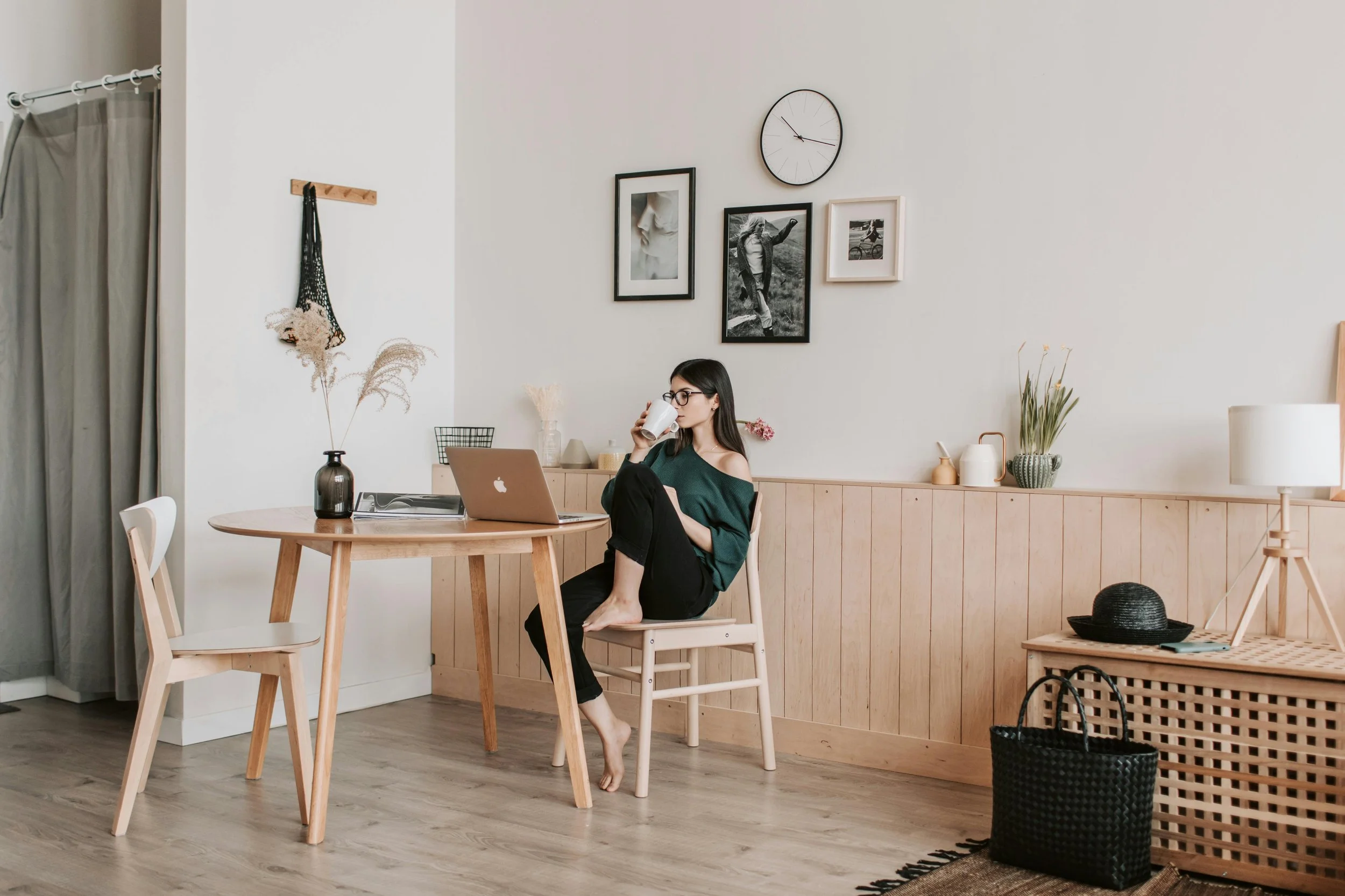 A woman with glasses and long dark hair sitting barefoot at a wooden dining table, drinking from a mug, with a laptop open in front of her, in a minimalist modern kitchen or dining area with light wood accents, framed black and white photos on the wall, and various decorative plants.