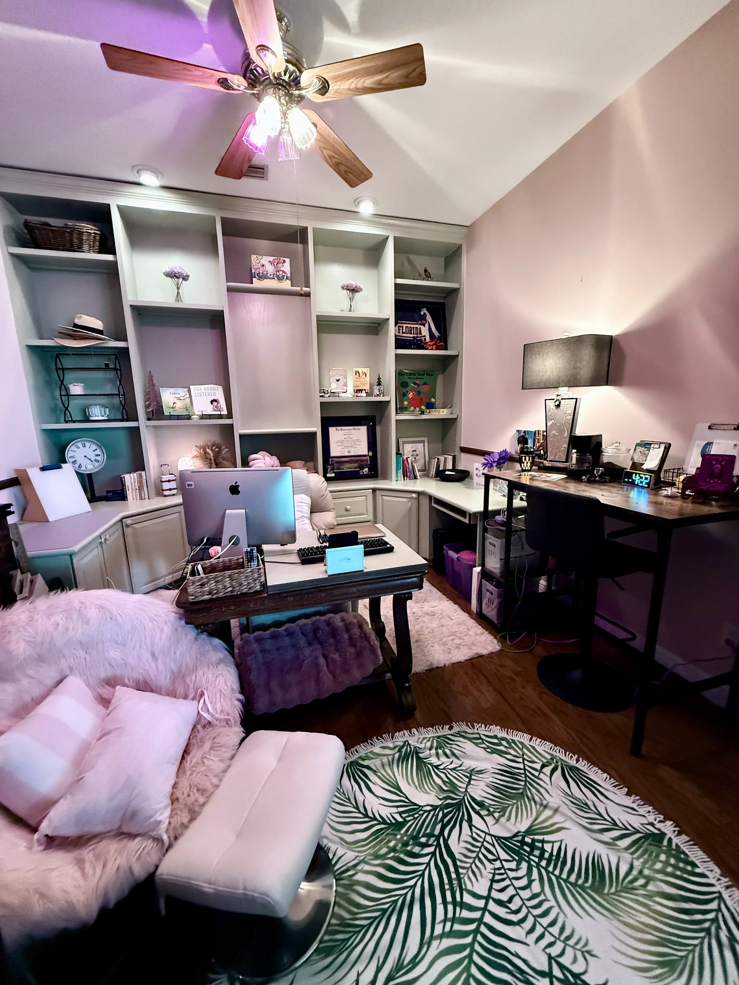 A home office with a white desk, a pink fluffy sofa, and a green leaf-patterned rug. There is a large white bookshelf with decorative items and books behind the desk. A ceiling fan with wooden blades and decorative lights is overhead. The walls are painted light pink, and there is a black table lamp on a side table with framed photos, books, and a digital clock displaying 4:22.