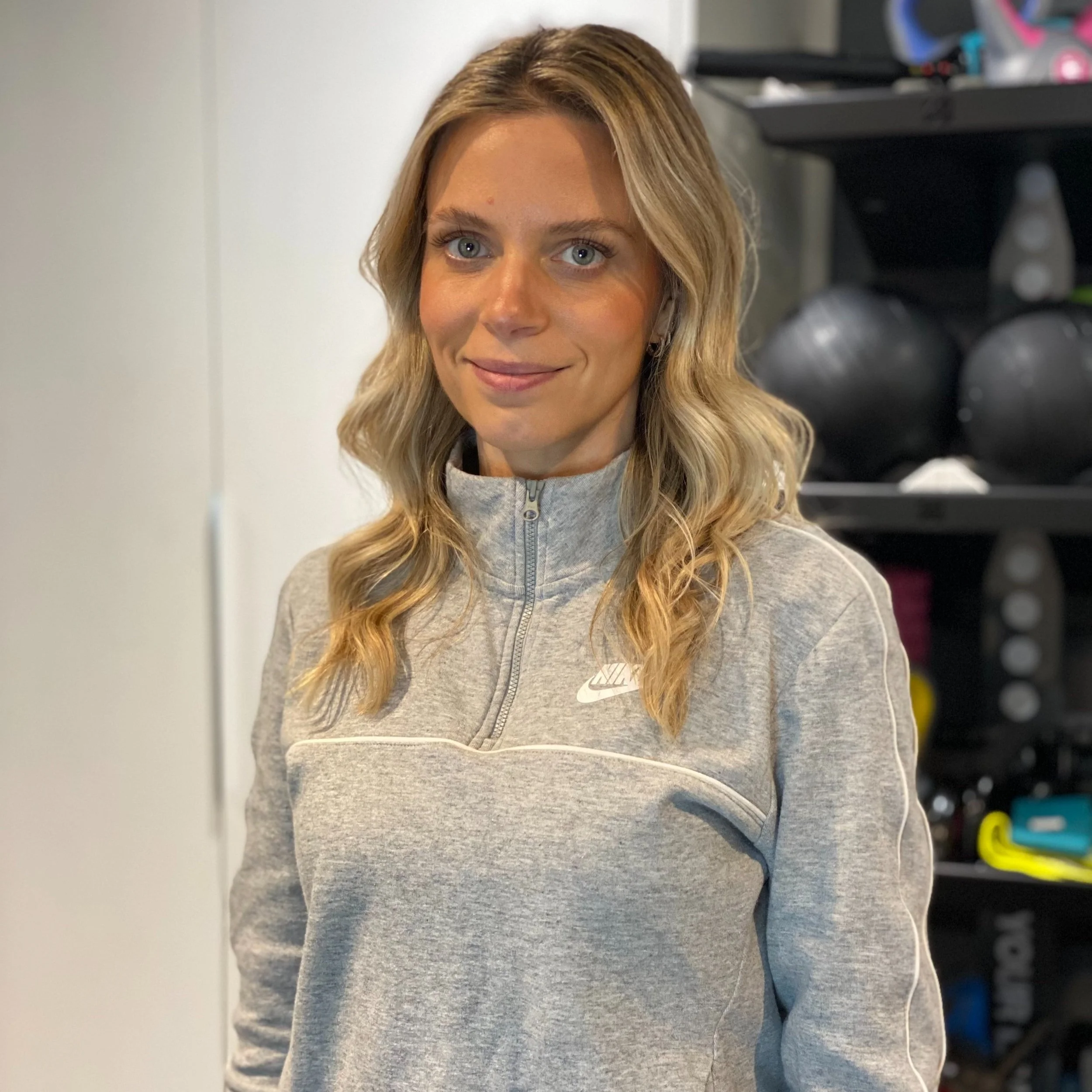 A woman with blonde, wavy hair and blue eyes smiling in a gym, wearing a gray Nike zip-up athletic jacket.