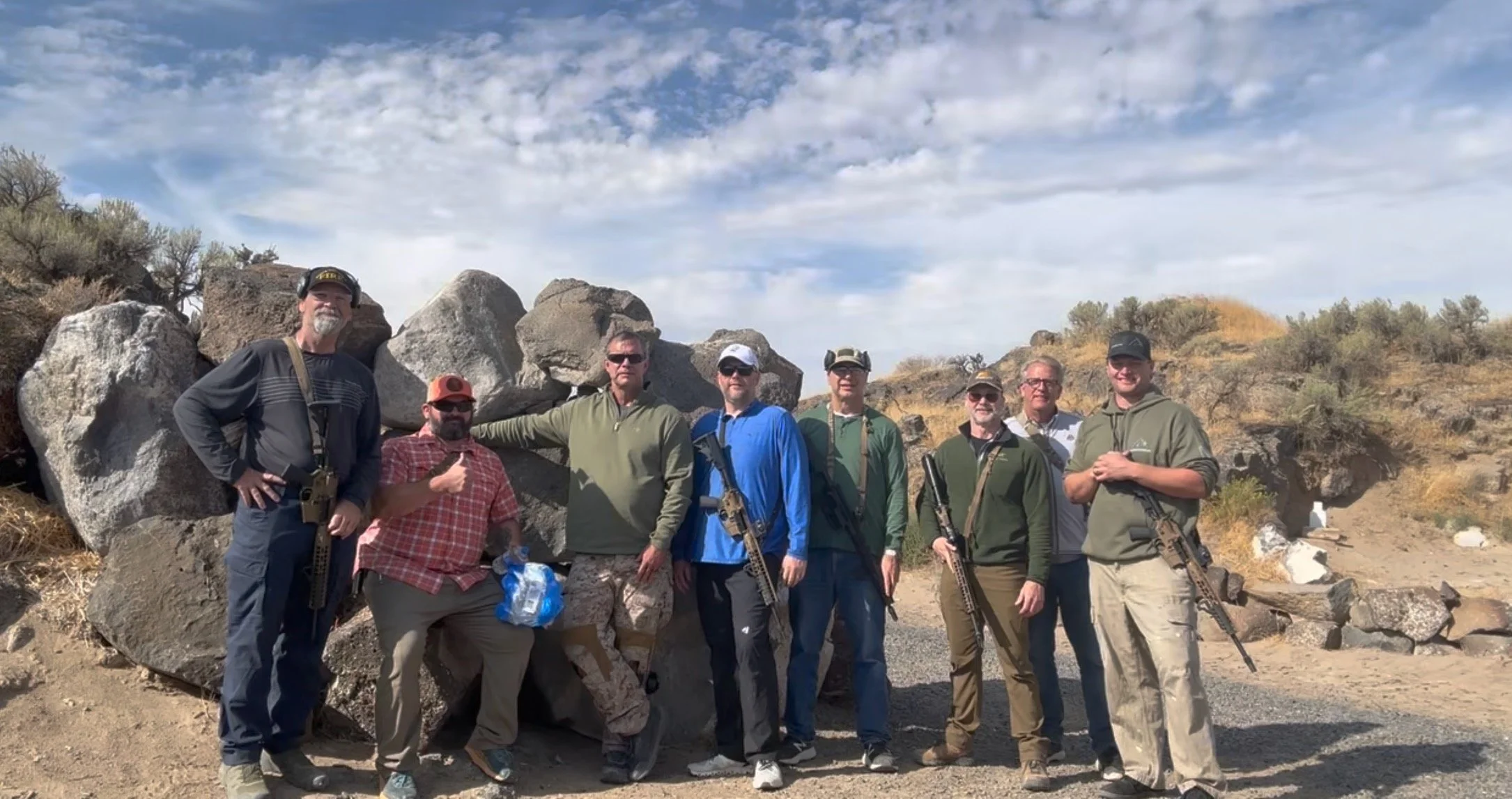 A group of eight men standing outdoors in front of rocks and dry bushes, some holding rifles, dressed in casual outdoor clothing, with a partly cloudy sky above.