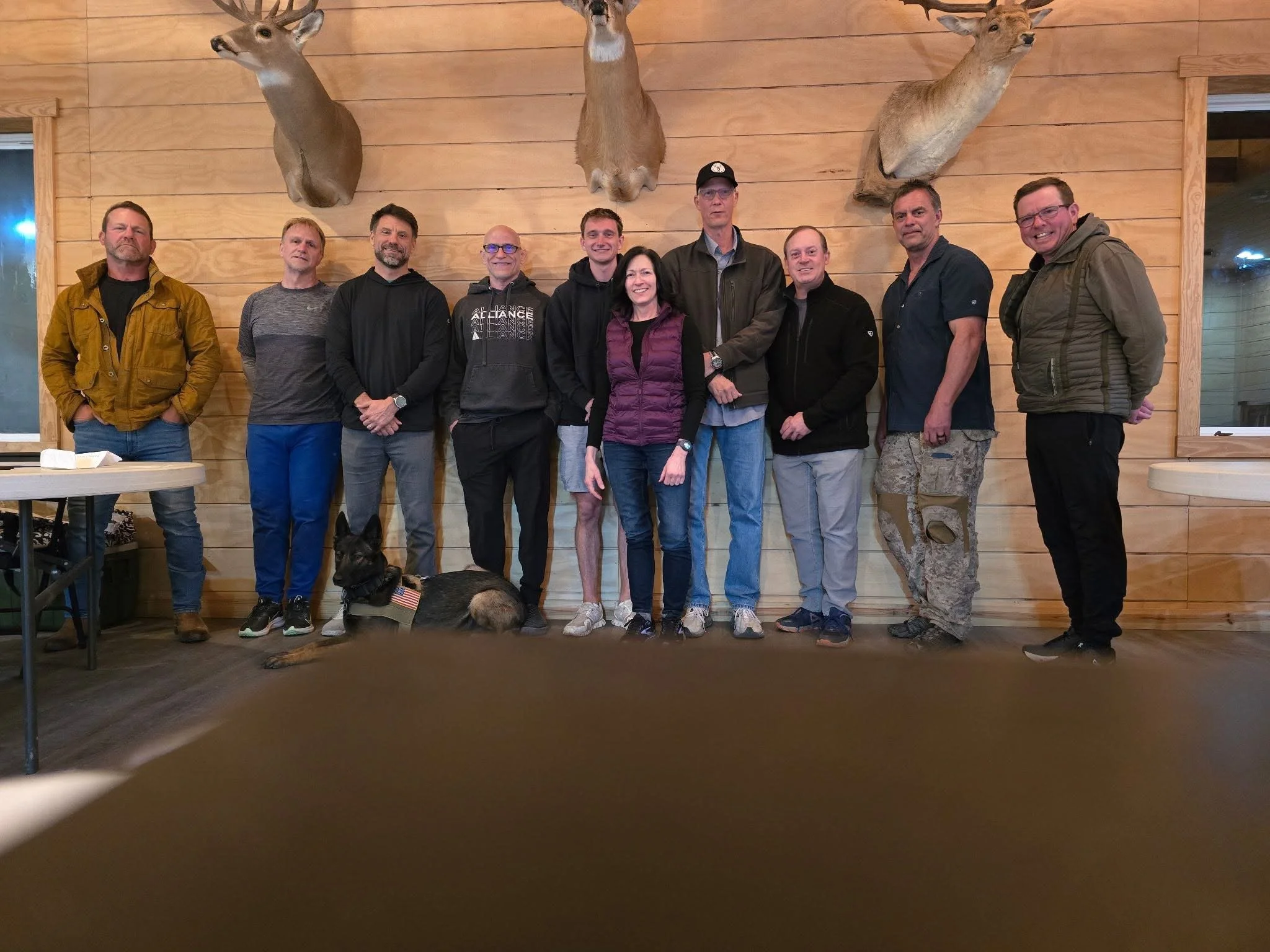 A group of ten people standing in front of a wooden wall with three mounted deer heads above them, and a dog lying on the floor in the foreground.