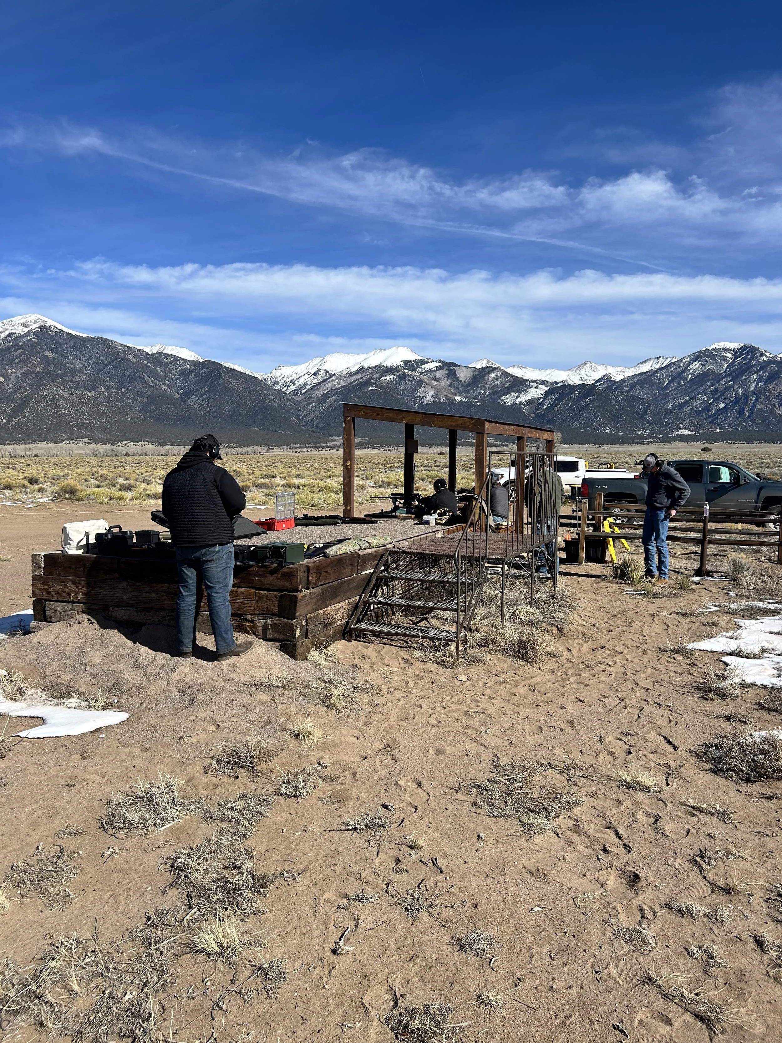 People preparing shooting equipment in an outdoor desert area with mountains and snow in the background under a blue sky with scattered clouds.