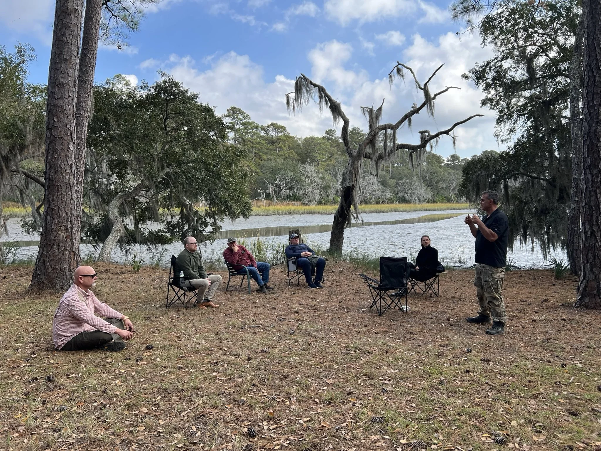 A group of people gathered outdoors near a river, with some sitting on chairs and a man standing while speaking. The setting is a wooded area with tall trees and Spanish moss hanging from the branches. The sky is partly cloudy.