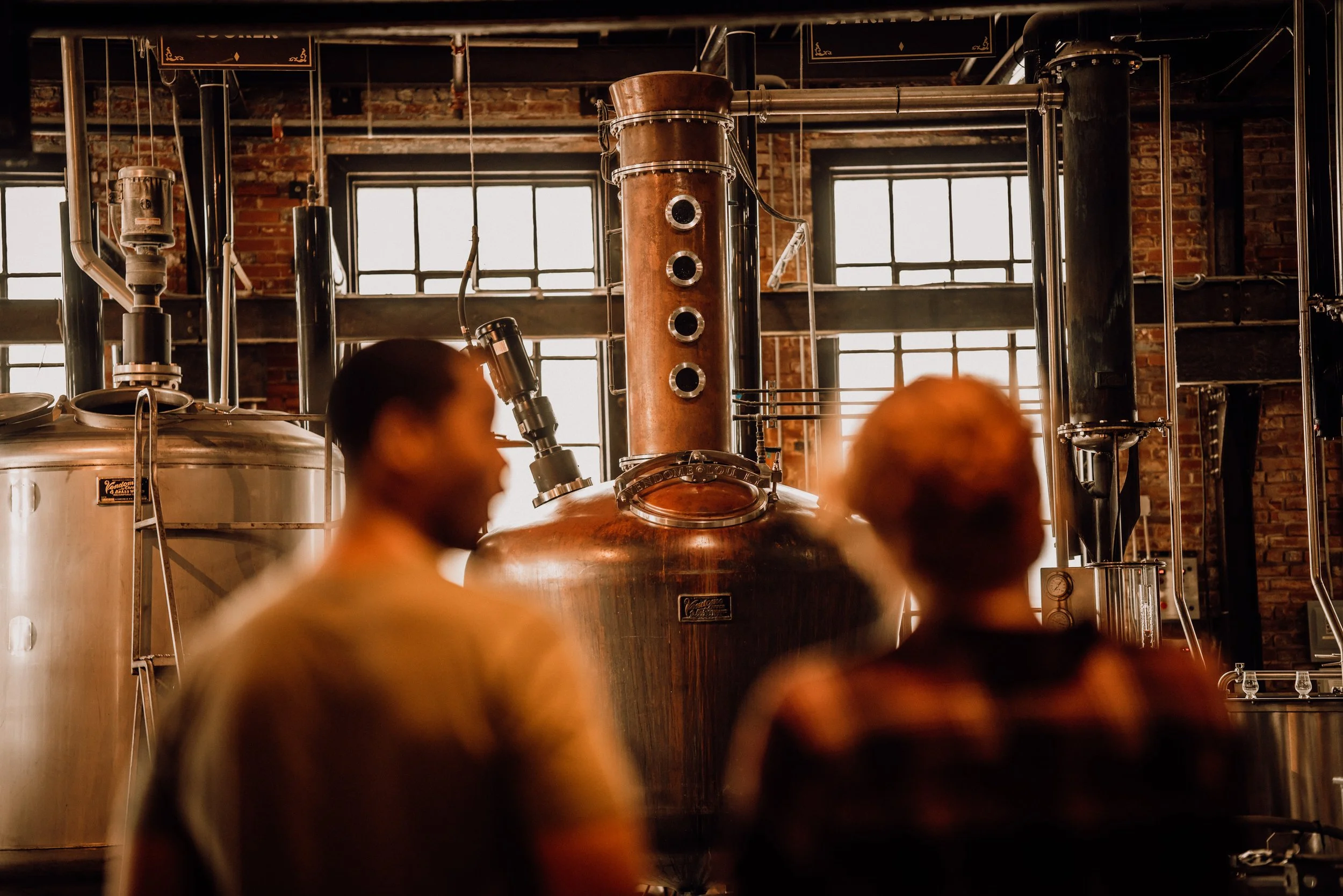 Two guests on distillery production floor looking at large copper still