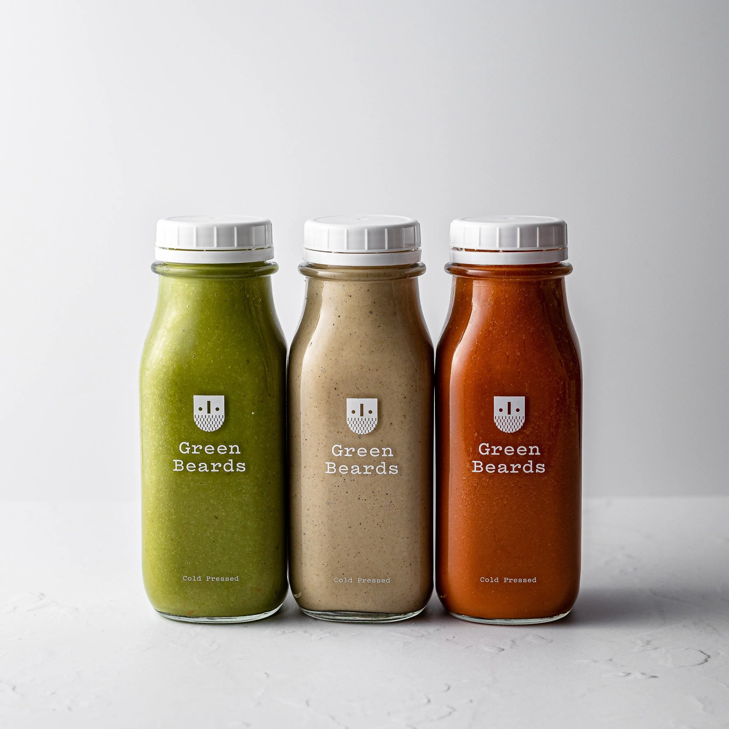 Three glass bottles of different colored sauces, with white caps, labeled 'Green Beards,' 'Green Beards,' and 'Green Beards,' from left to right, on a white background.