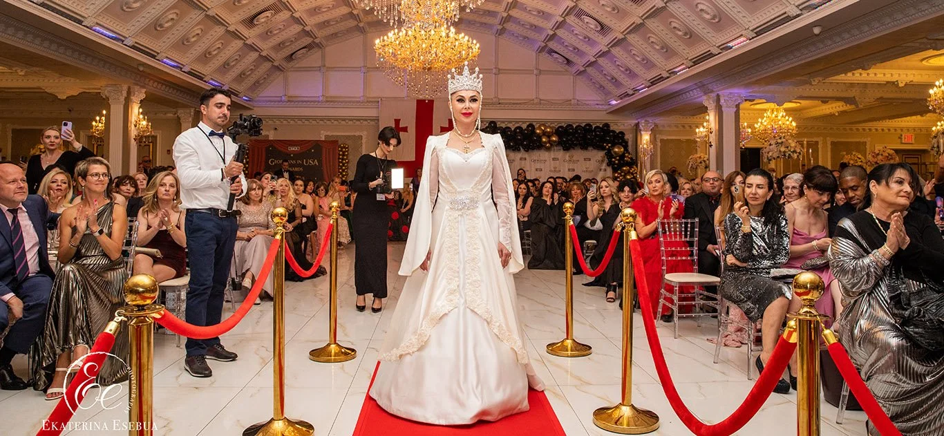 A woman in a white wedding gown with a crown and cape walking on a red carpet at a wedding reception or event, with seated guests clapping and watching the ceremony in a grand decorated hall.