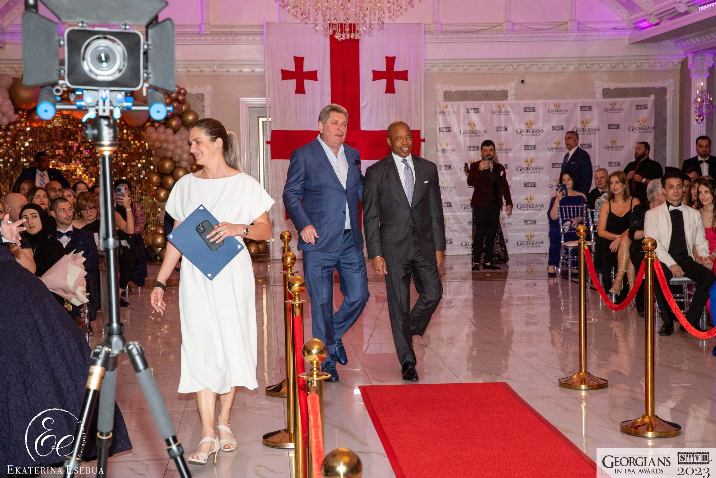 A woman in a white dress walking down a red carpet at a formal event with a large Georgian flag in the background. Several people are seated on either side, and there are event banners and balloons as decorations.