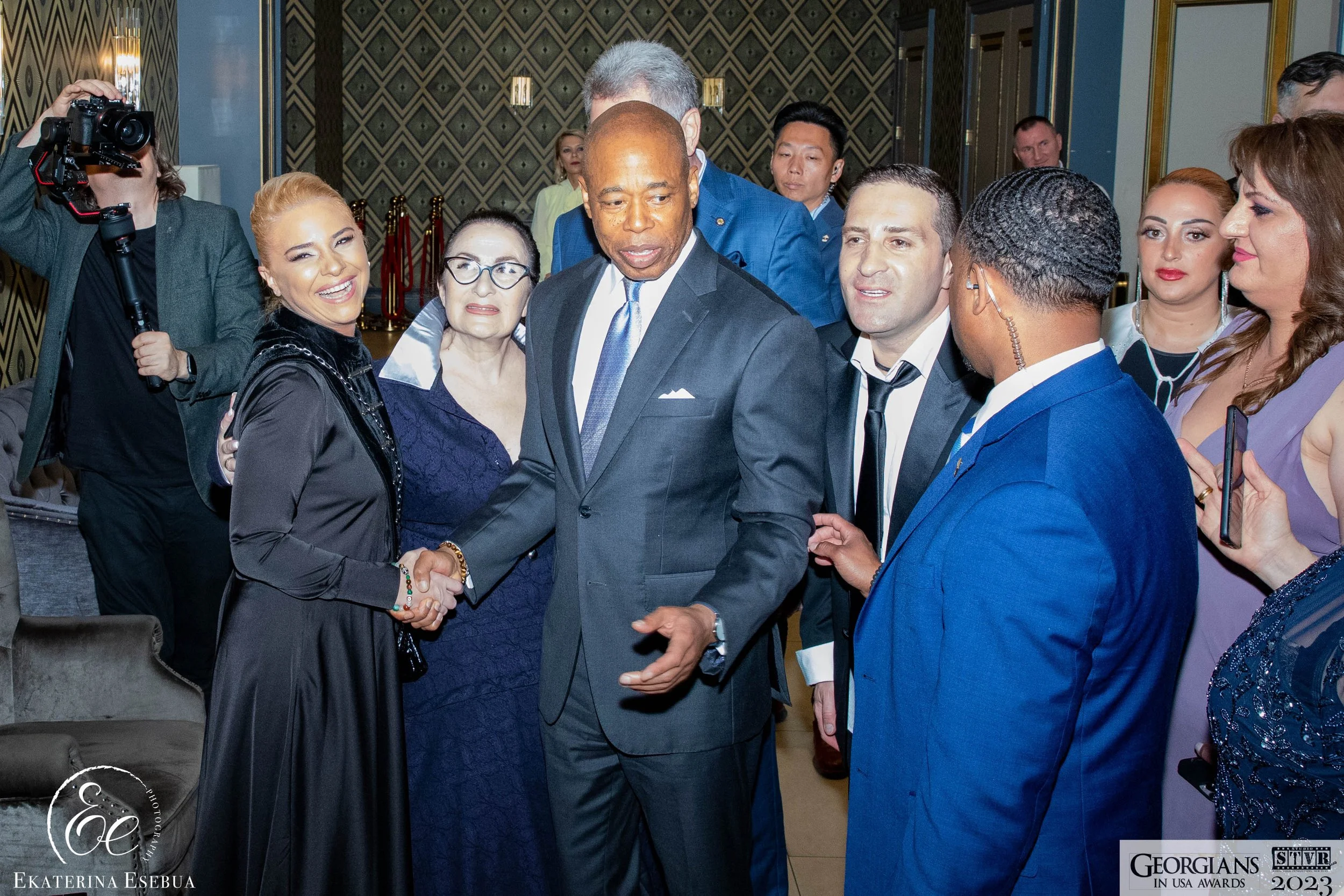 A man in a black suit shaking hands with a woman in a dark blue dress at an event, surrounded by other well-dressed people, with a woman taking a photo on the left.