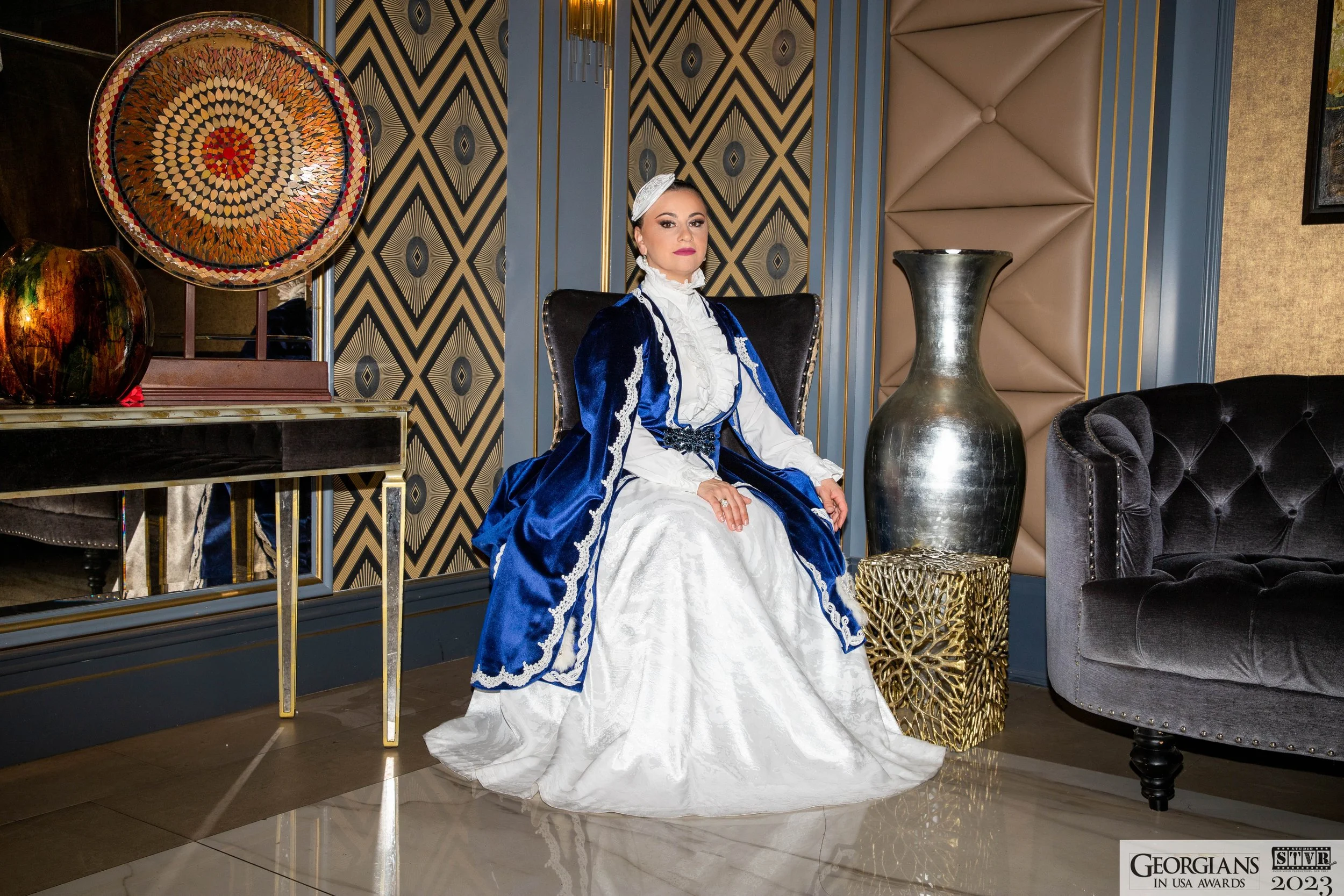 A woman dressed in a historical style white gown with a blue velvet cloak featuring lace trim, seated in an elegant room with decorated wallpaper, a large decorative plate, and gold accents.