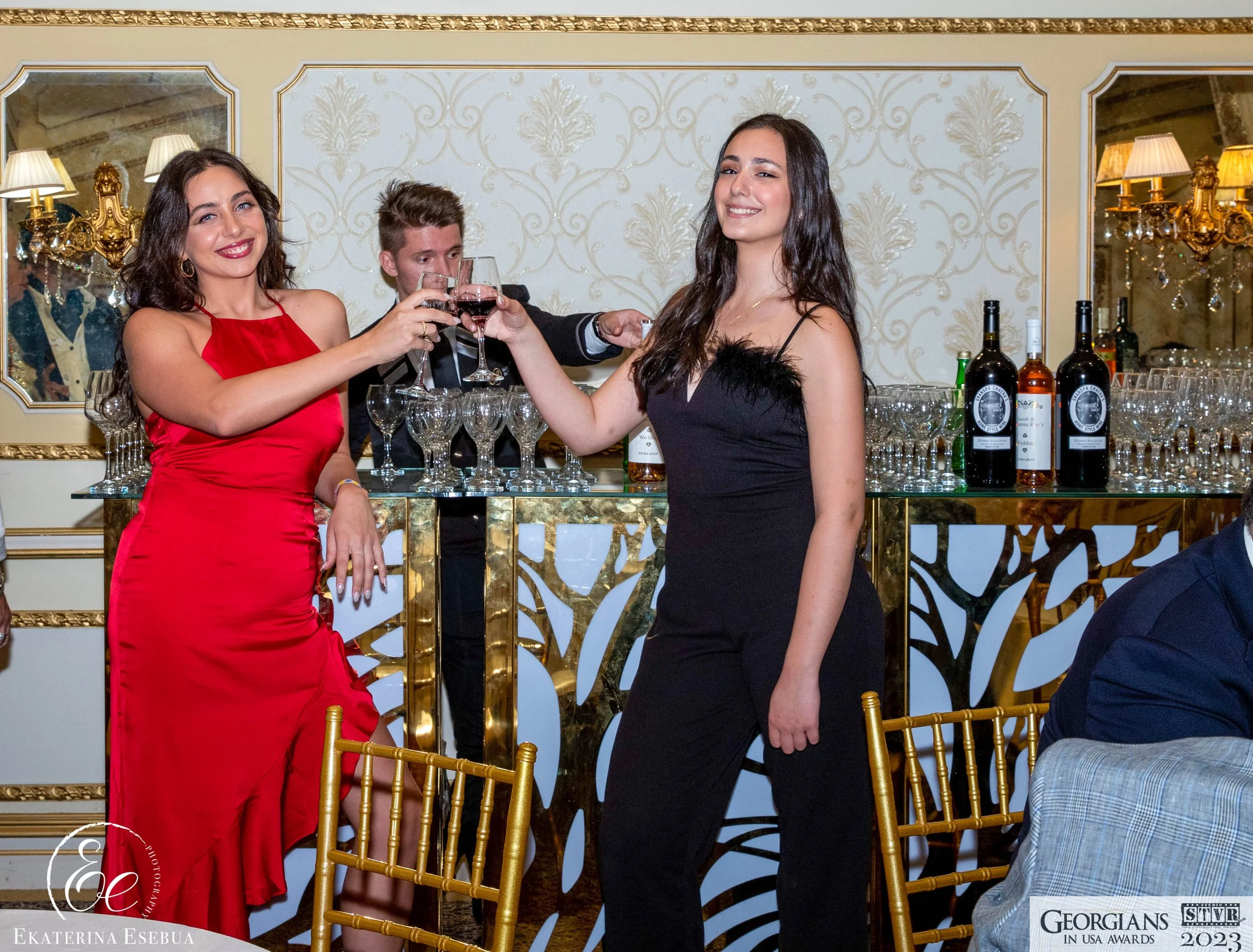 Two women in elegant dresses clink glasses of red wine at an event, smiling, with a bar behind them containing wine bottles and glasses, and ornate decor including gold accents and wall sconces.