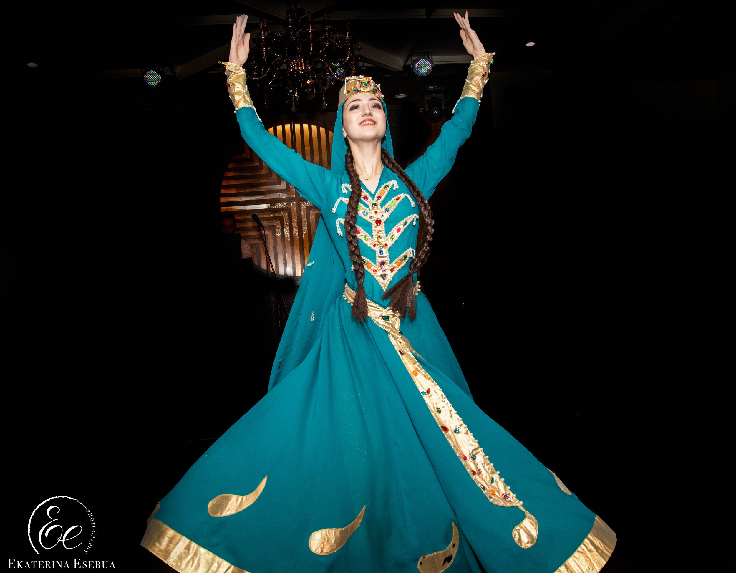 A woman dressed in traditional costume with a blue dress, gold accents, and a crown, raising her arms in a celebratory pose at an indoor event with a chandelier and stage lighting in the background.